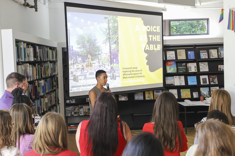 Photo of woman speaking to audience in front of screen that reads 