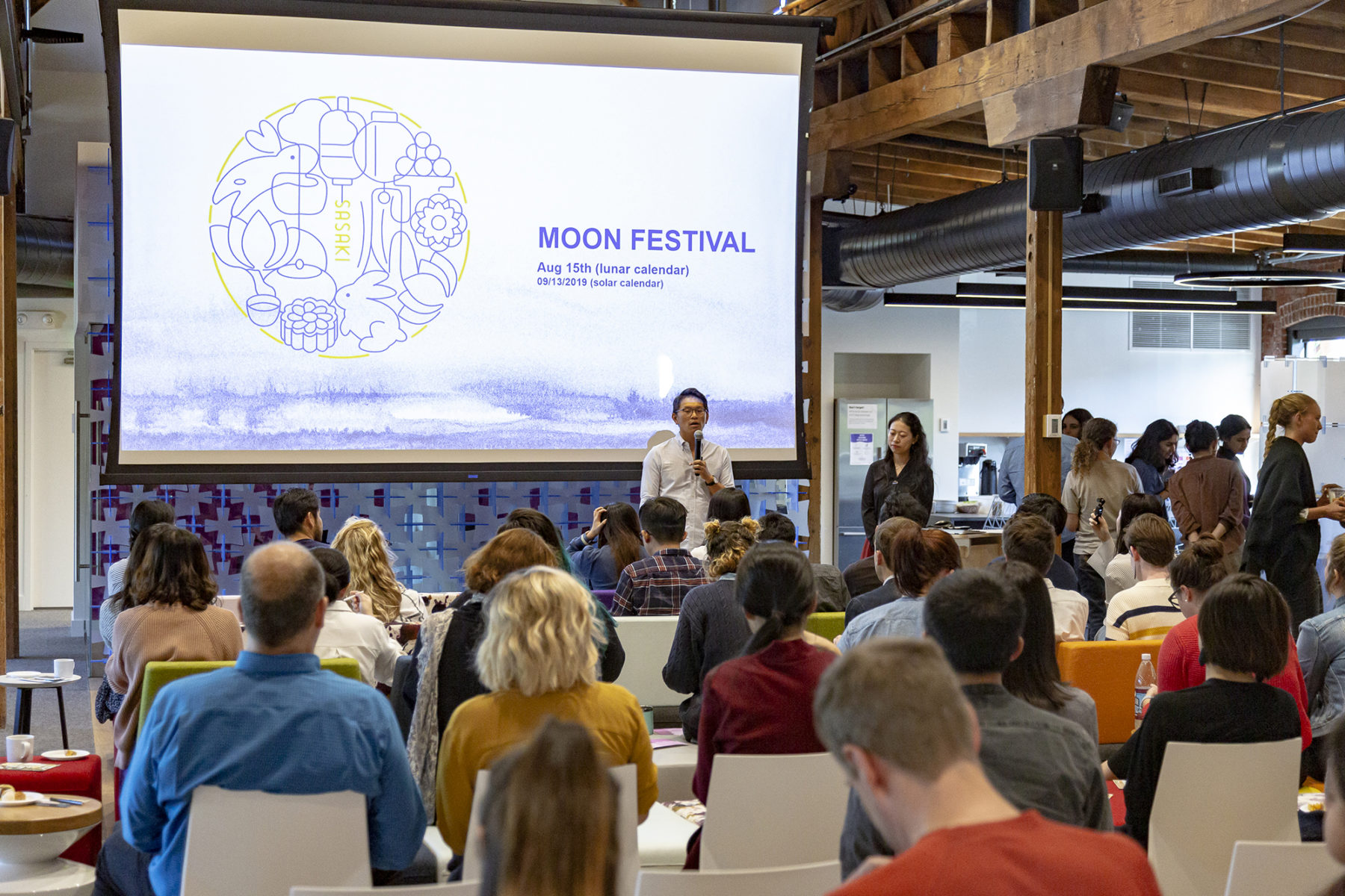 Photo of man addressing a group with Moon Festival graphic on large screen behind him