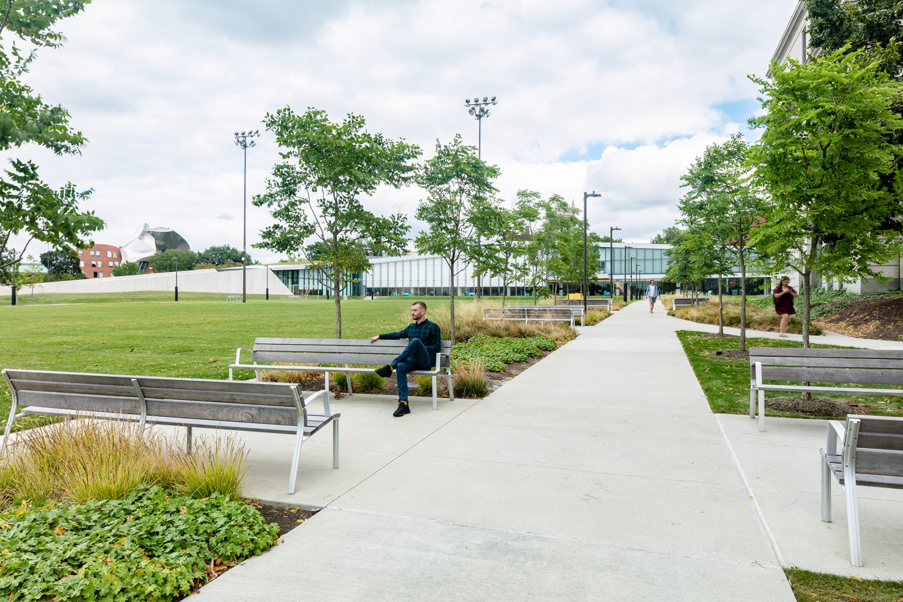 walkway towards large building, lawn on left, man sitting on bench