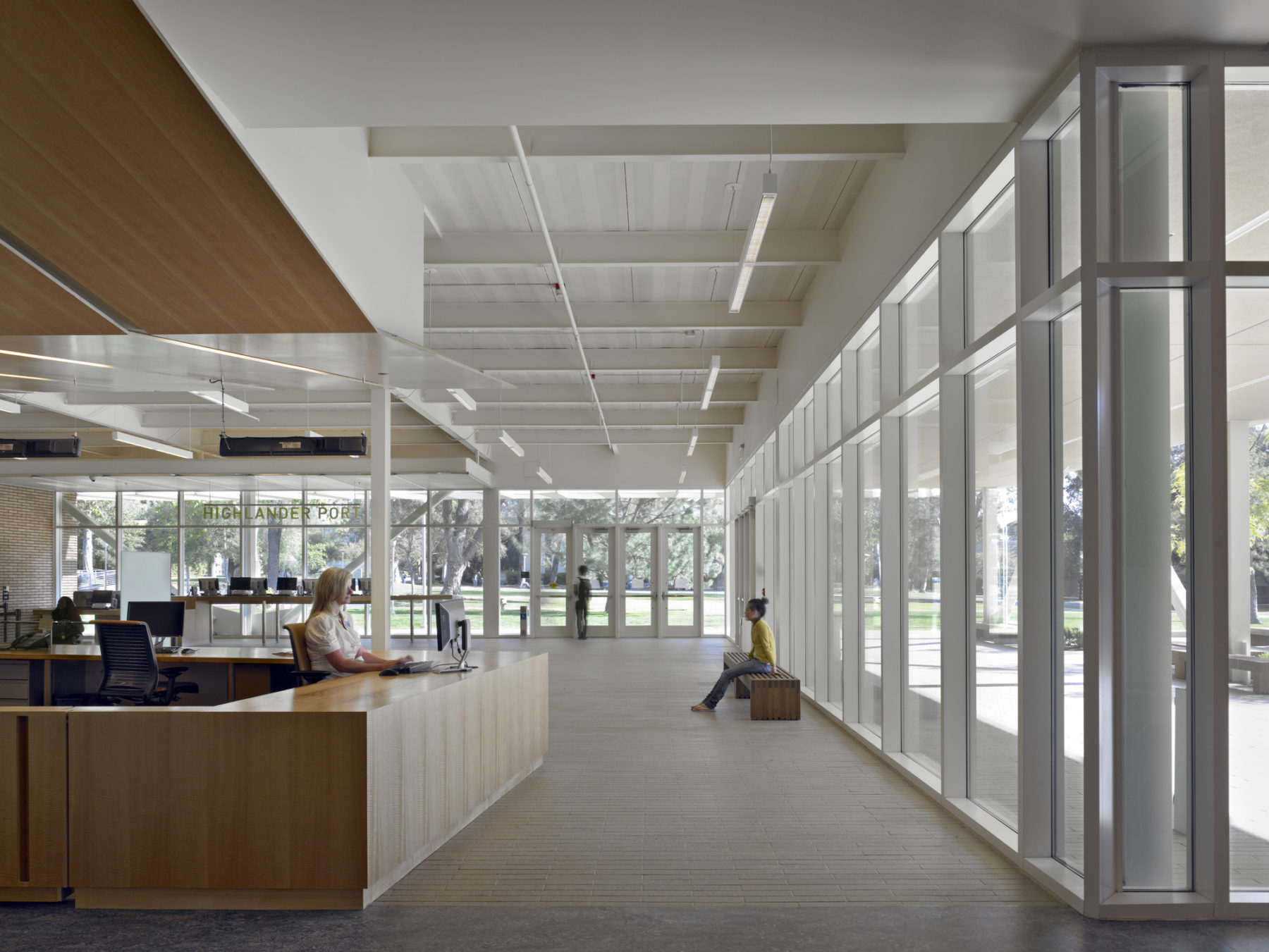 Interior photo of the building entrance and welcome desk. A student sits on a bench in the background