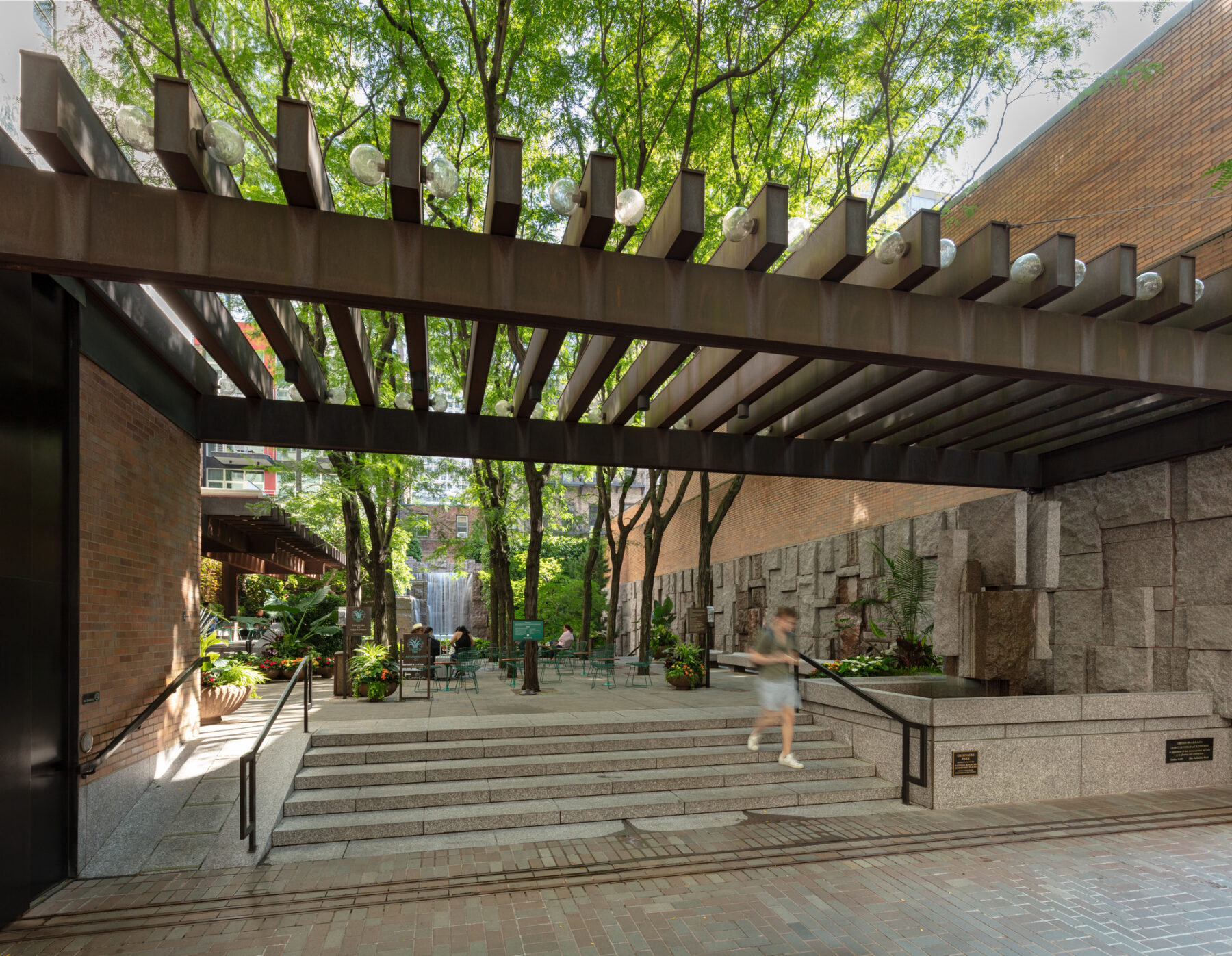 Photo with street view of the park entry, framed by overhanging trellis with trees growing behind and above and visitors walking in and out of the entry stairs.