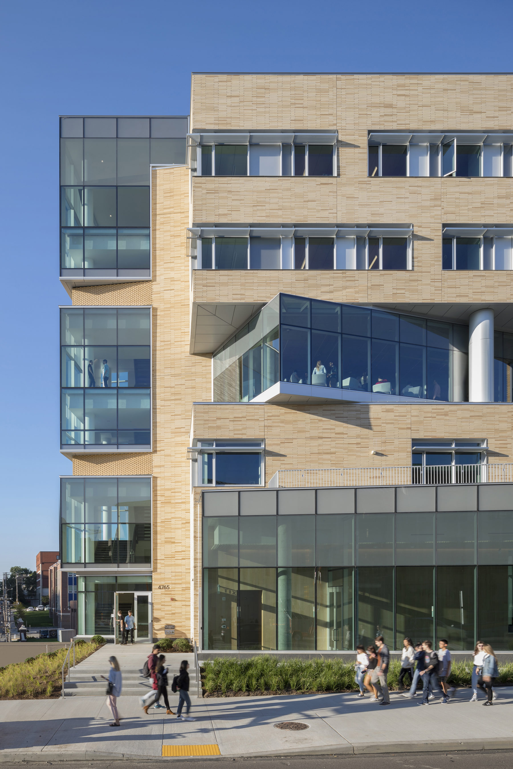 Exterior view of side of building with people walking, transparent windows from the building enabling us to see the activities inside.