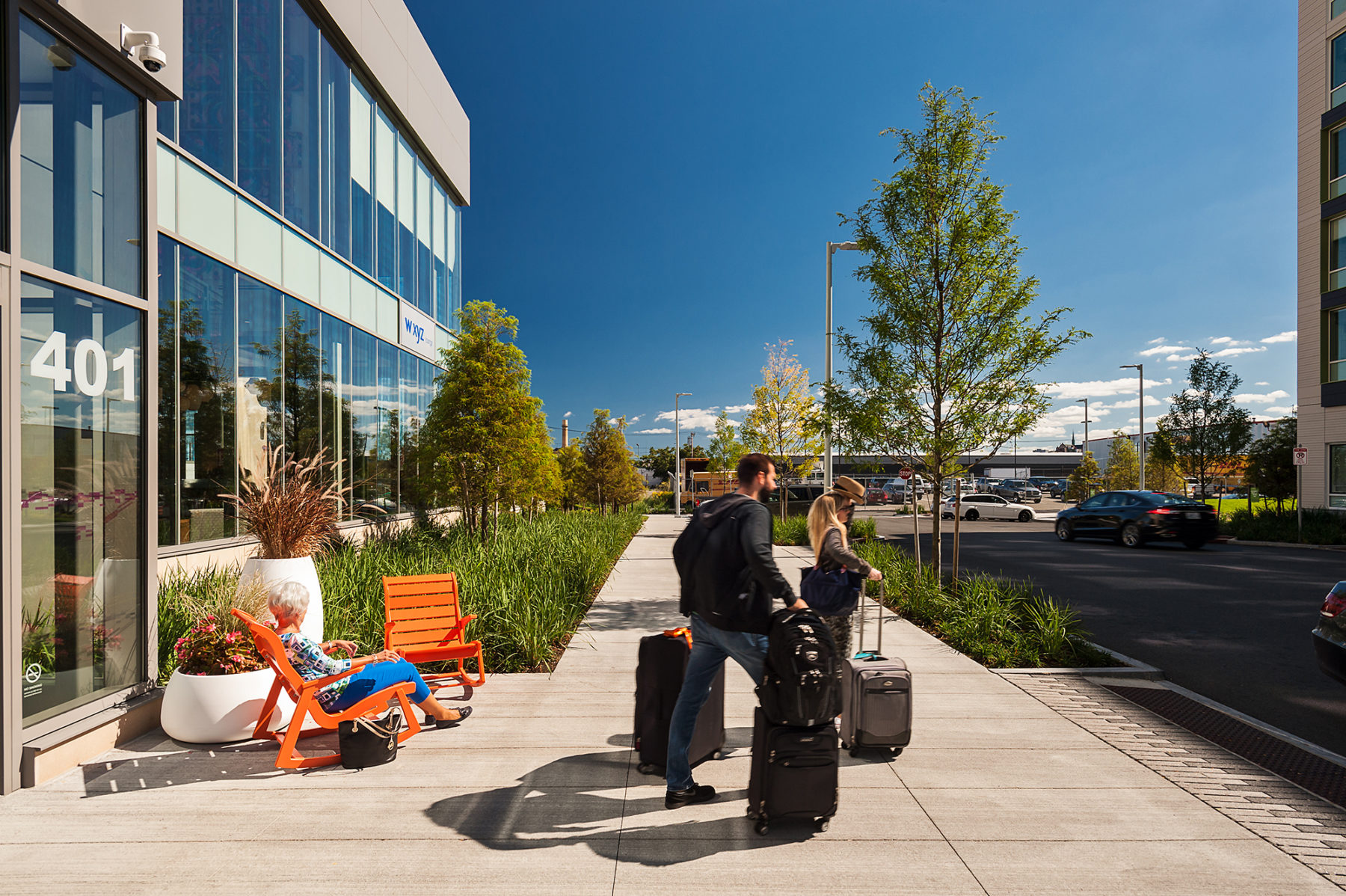 Photo of a couple exiting hotel with luggage. A woman sits in an orange chair on the sidewalk.