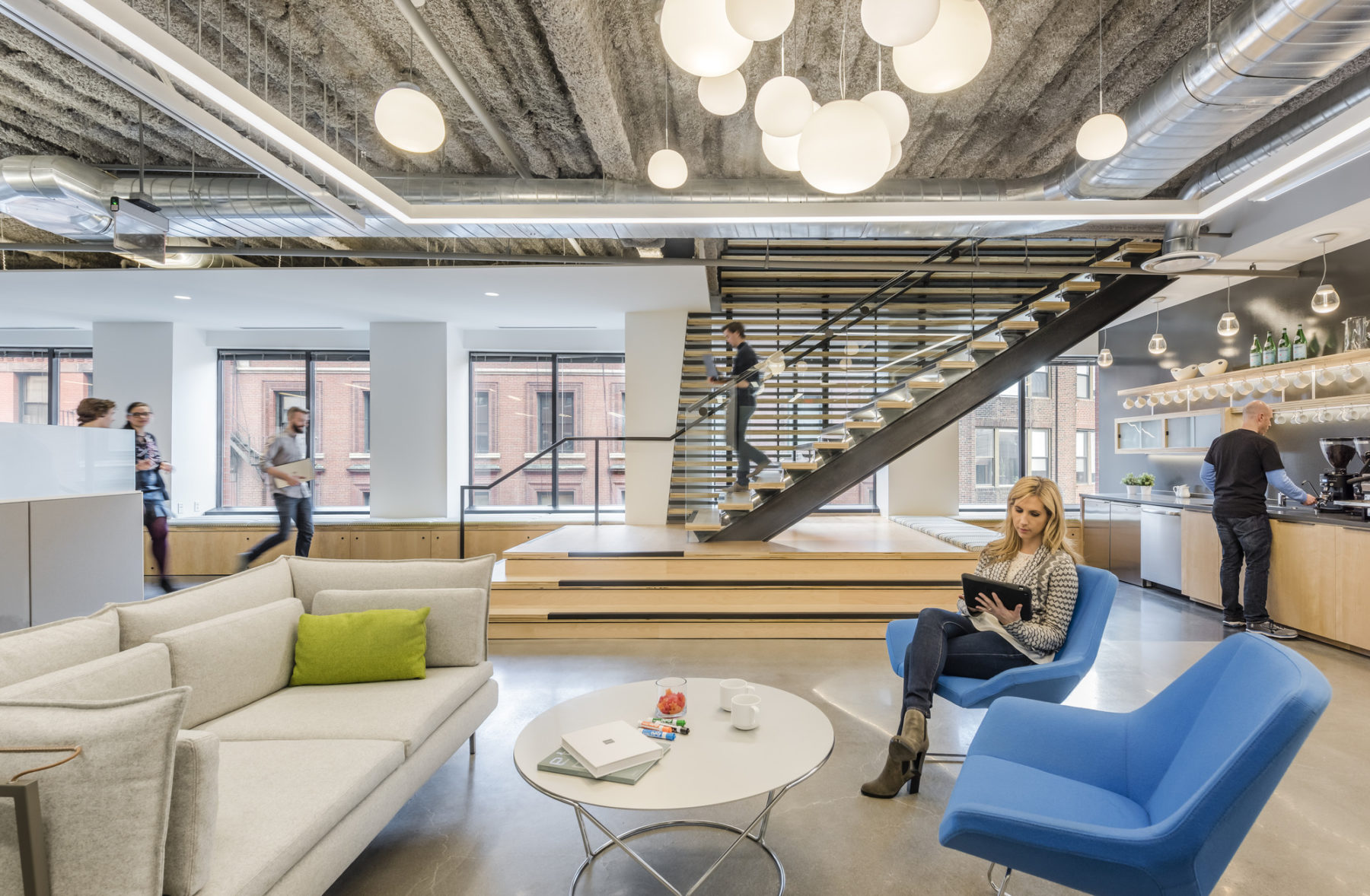 Open office space, people engaged in various forms of activities, woman sitting on sofa set at the front, kitchen at the right, and stair at the back. Bright lights, big windows.