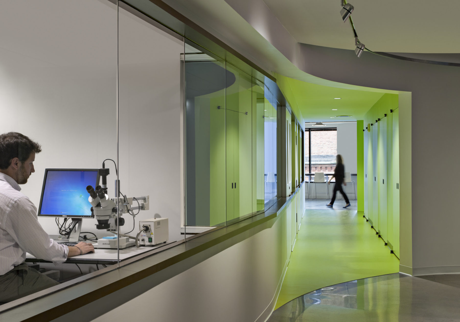 Man working on his desktop and microscope in a see-through office area towards the left, while we can look and see a greenly lit hallway on the right.
