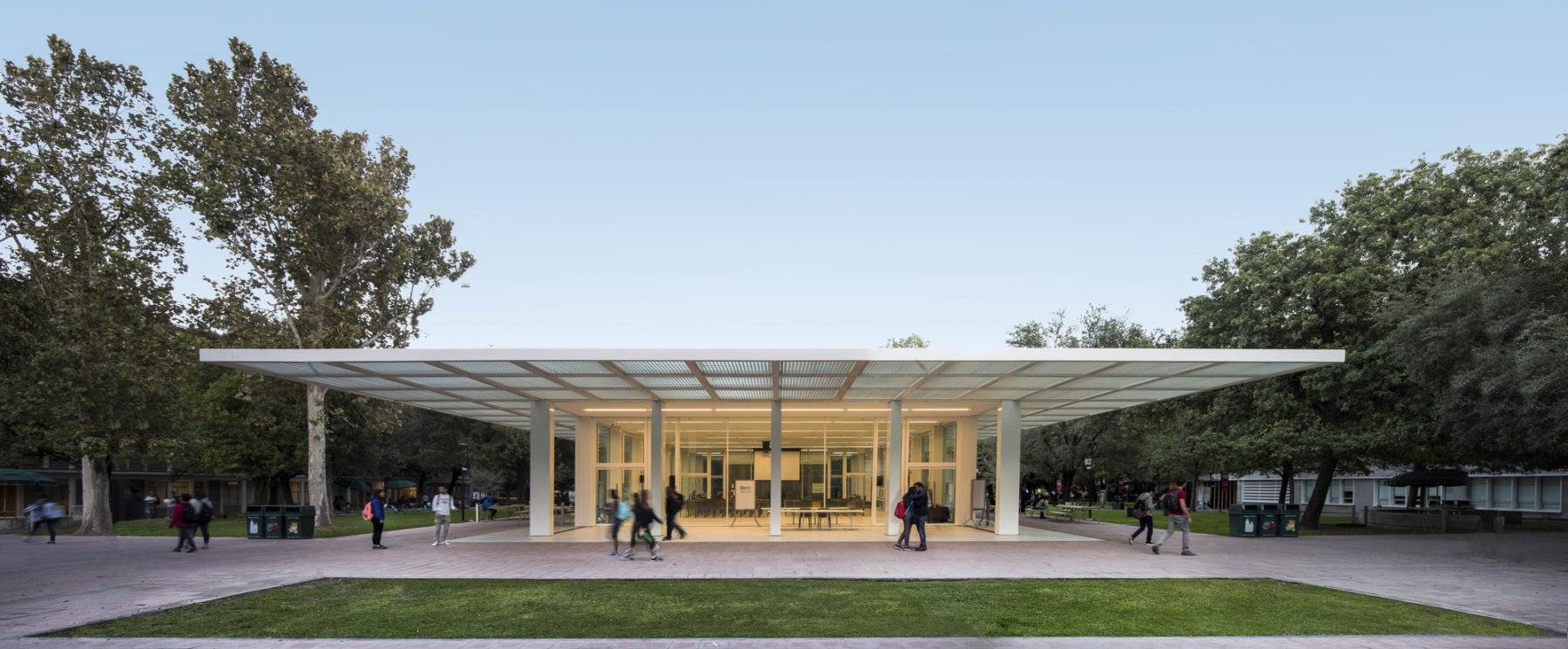Color photo of a compact, glass-enclosed pavilion sited on a lawn with a deep overhang for sun shading. students walk by