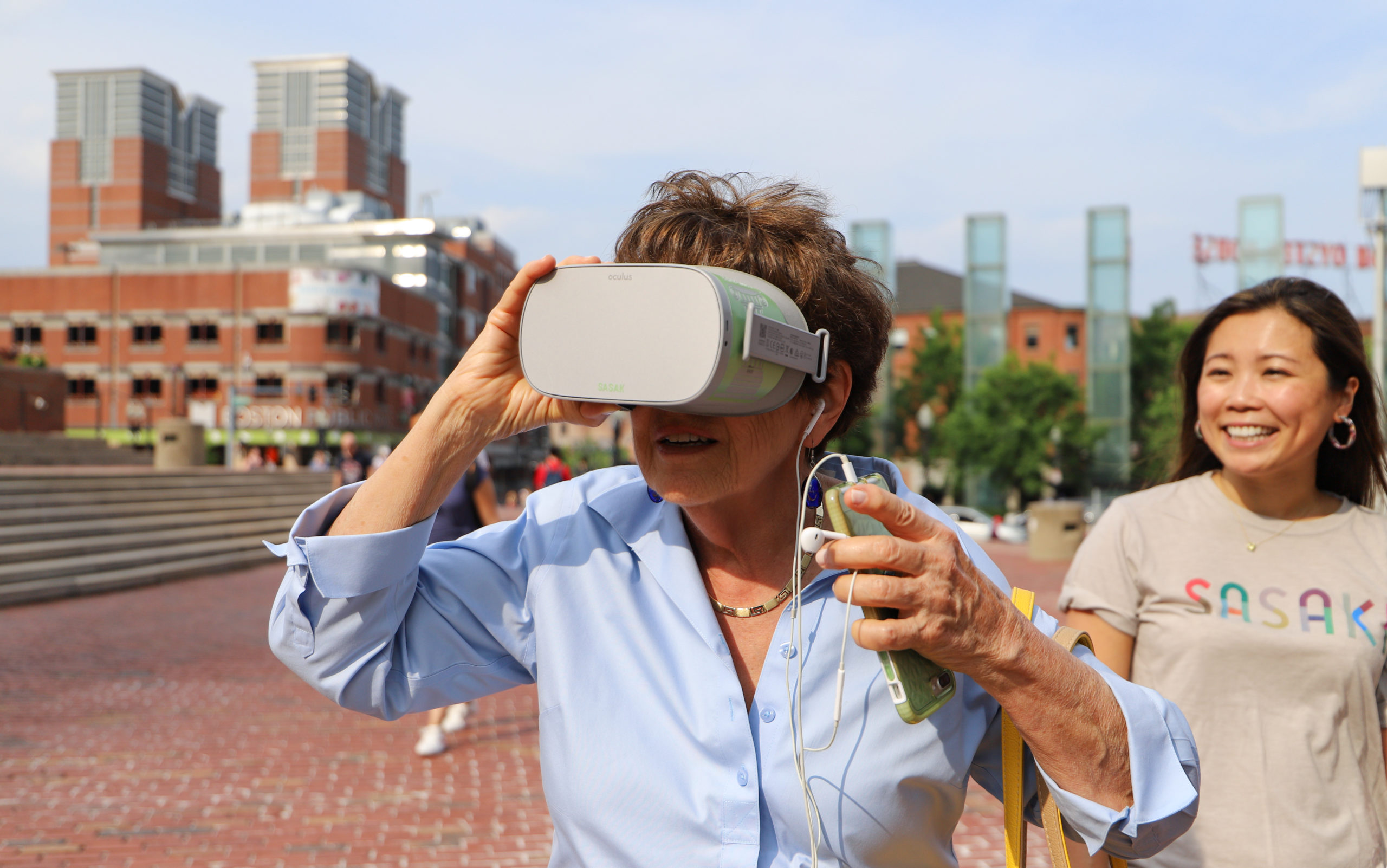 Older woman looking through VR goggles, person smiling in background