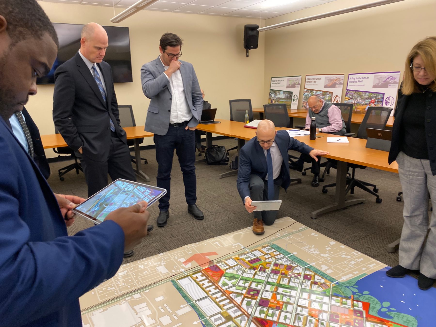 a group gathers around a large map placed on the floor. One participant looks at 3D content on a tablet device.