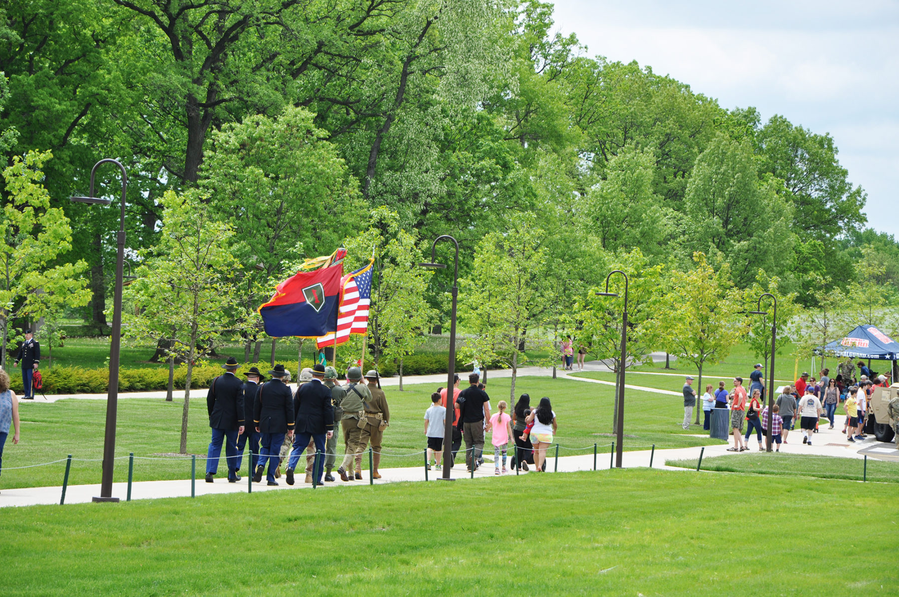people carry flags down a paved pathway surrounded by green grass fields