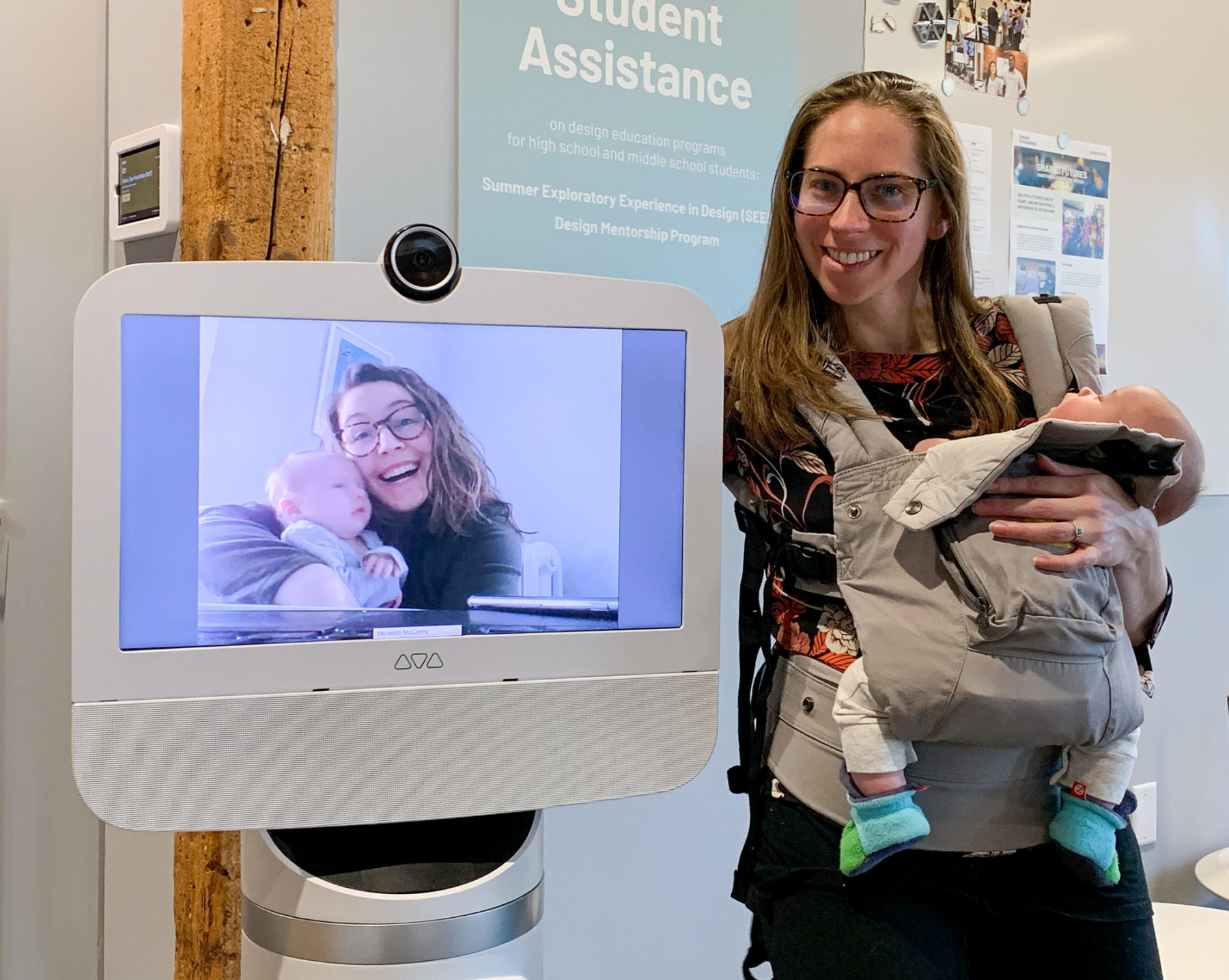 Woman holds baby and poses next to a robot's screen showing another woman holding her baby