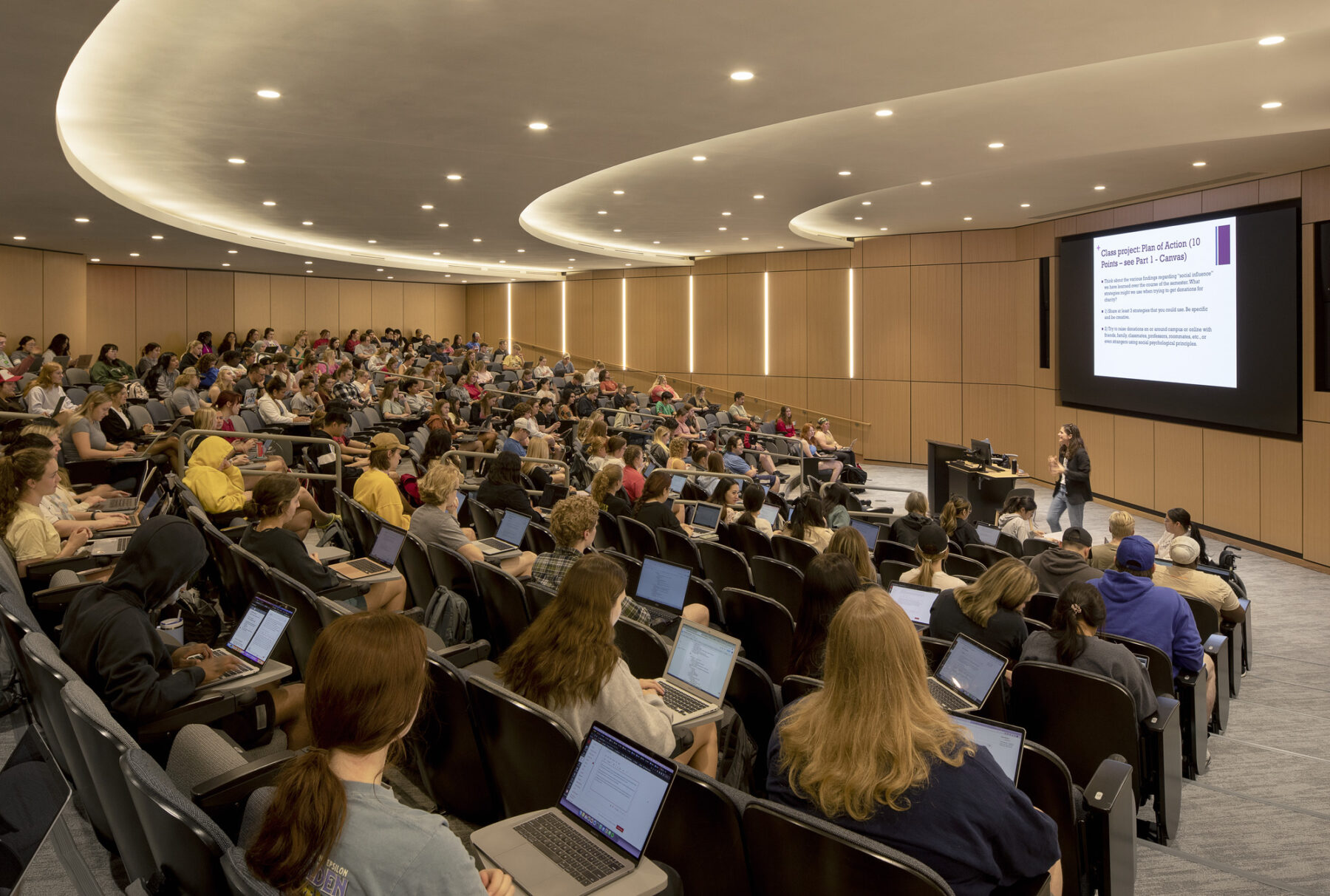 Photo of lecture hall filled with students taking notes on laptops and faculty presenting in front