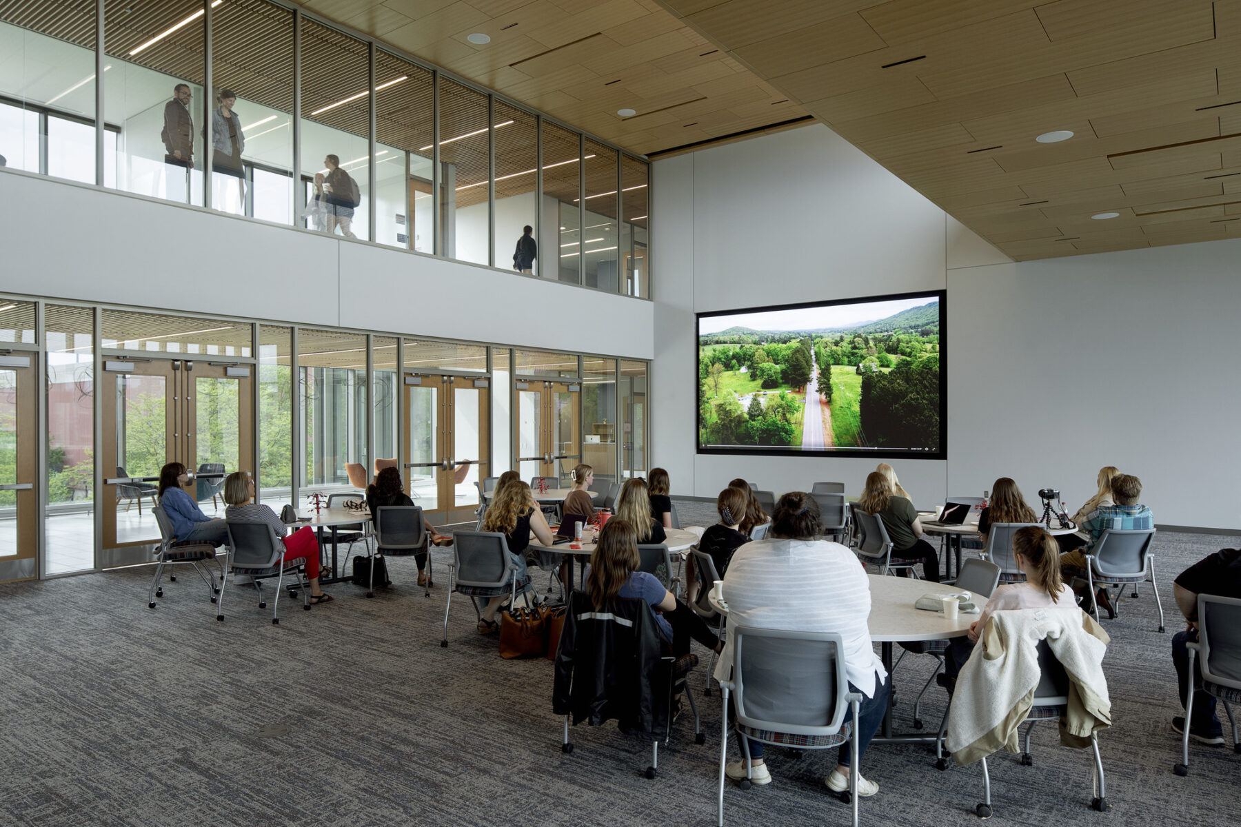 Photo of class presentations in an open, conference room space with large TV screen and windows with faculty and students looking in from the glass windows on the bridge above