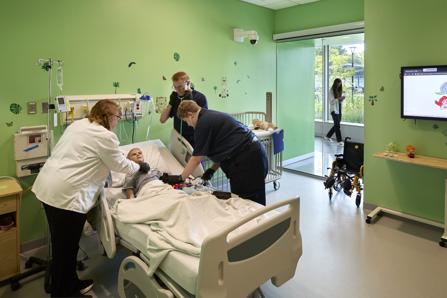 Nursing professor and two students during a pediatric simulation with a mannequin lying in a hospital bed.