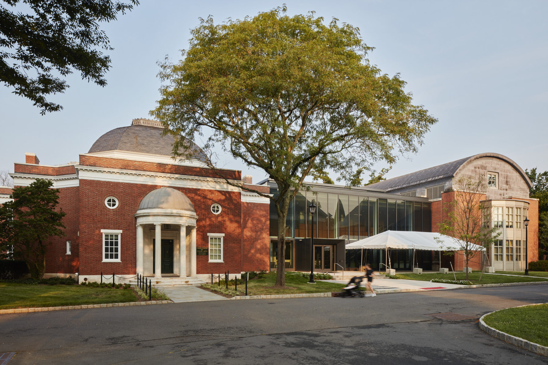 exterior photo of building with a large tree in front of the addition. A woman pushing a stroller walks in the foreground