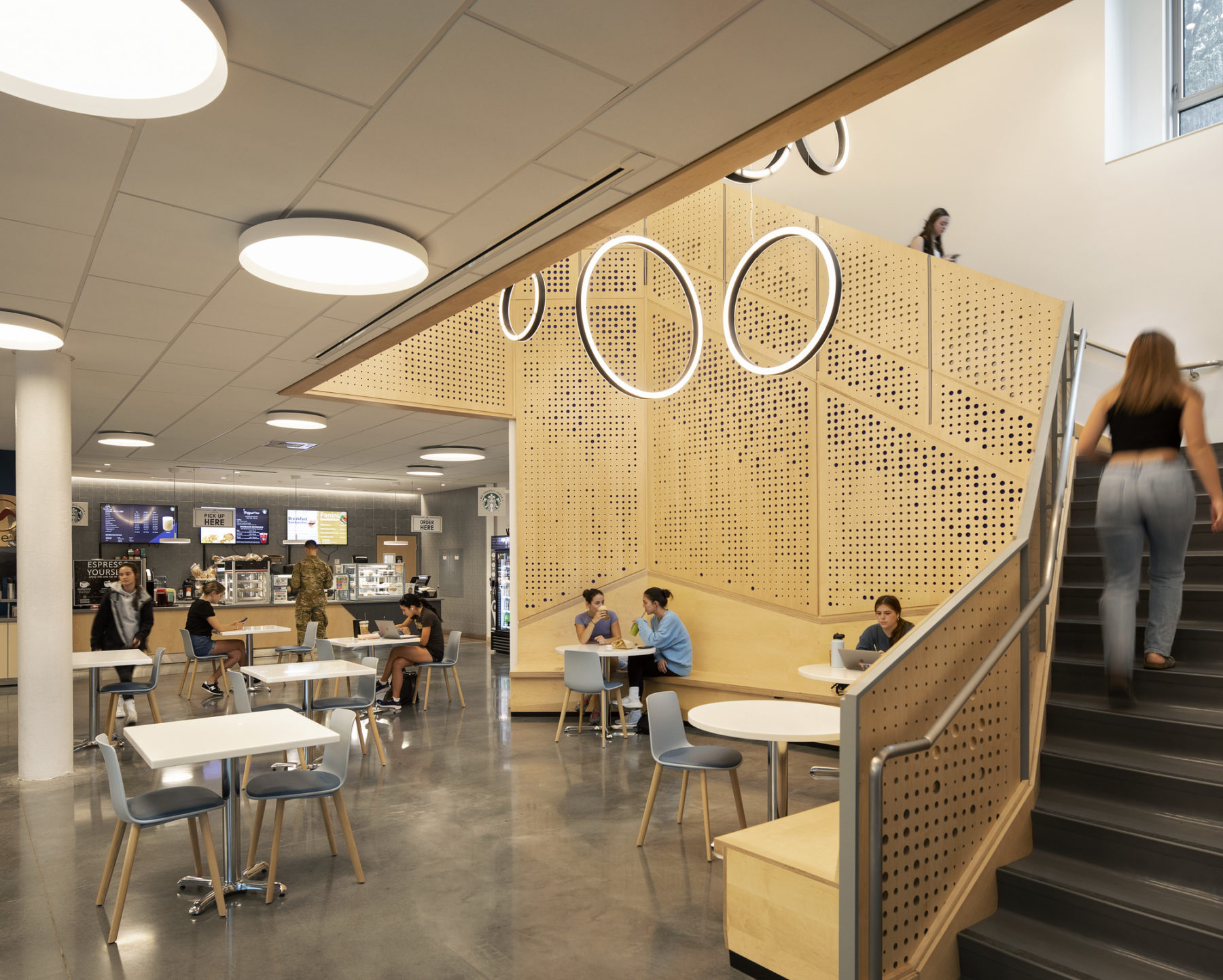interior photo of building cafe. Students sit underneath a circular light fixture suspended in stairwell