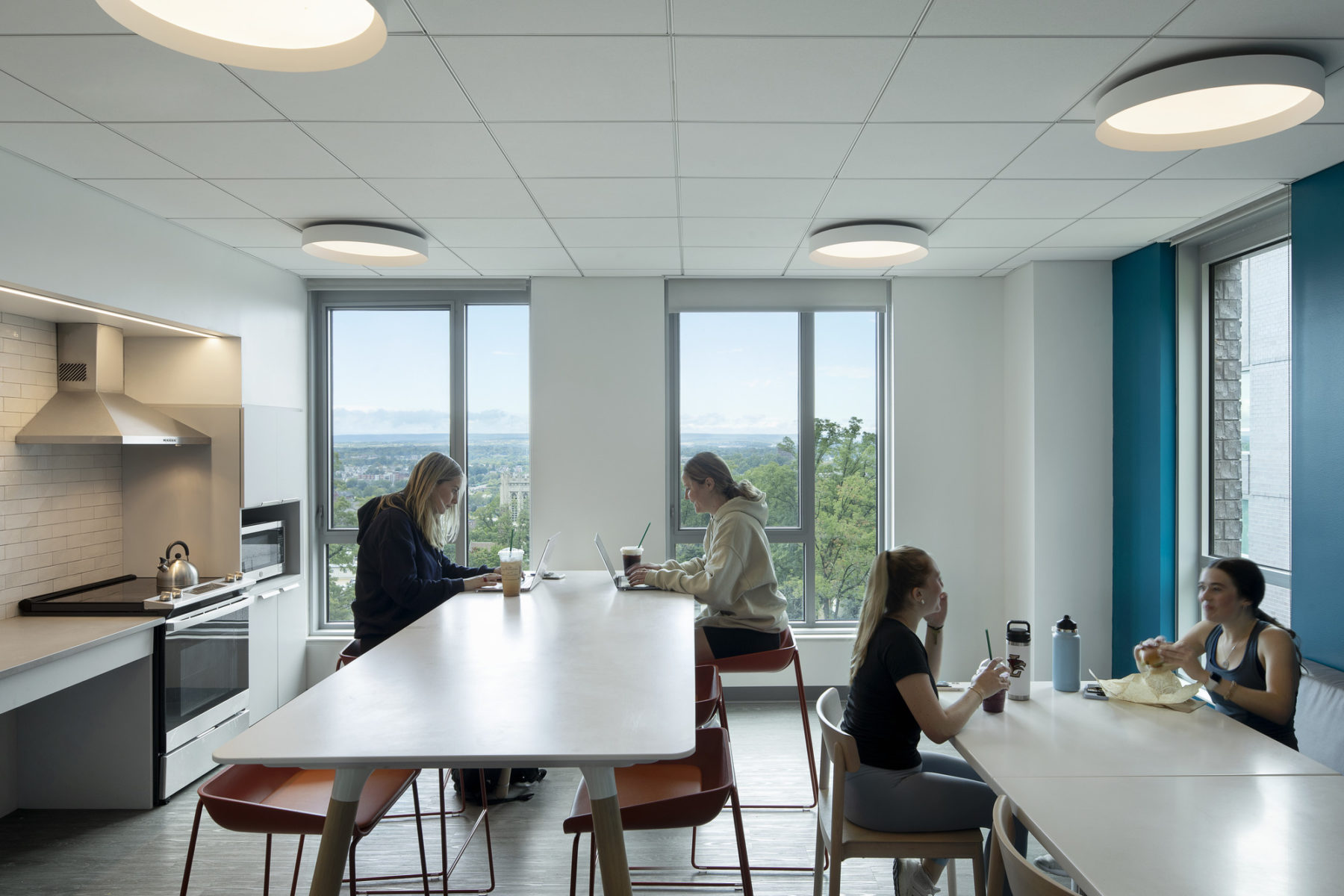 interior photo of students sitting at two tables in shared kitchen. two large windows overlook the campus in the background