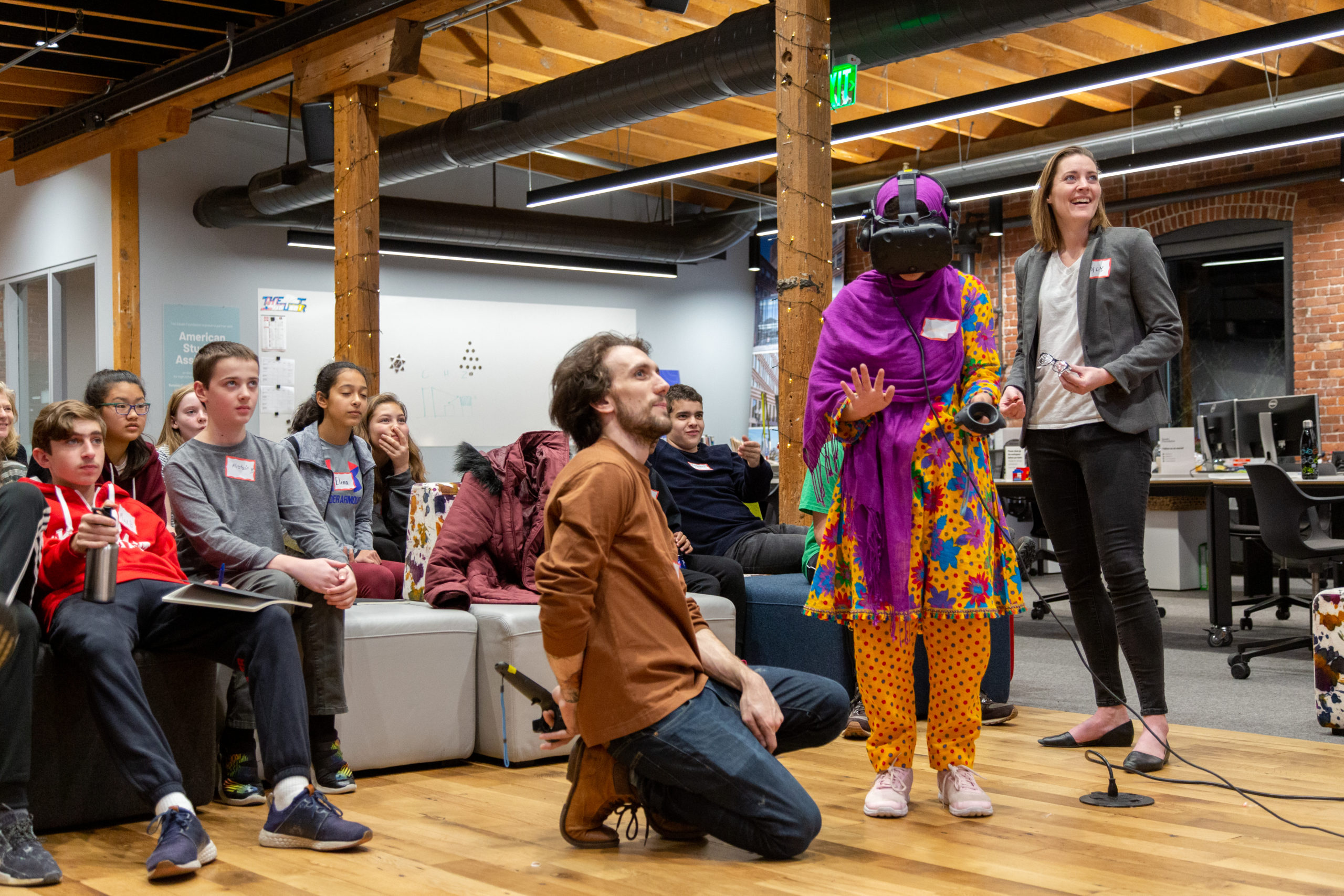 Emily, the author, smiling, stands next two a teenager wearing a VR headset. Another employee kneels beside the teenager to show her how to use the headset. In the background other teens look on