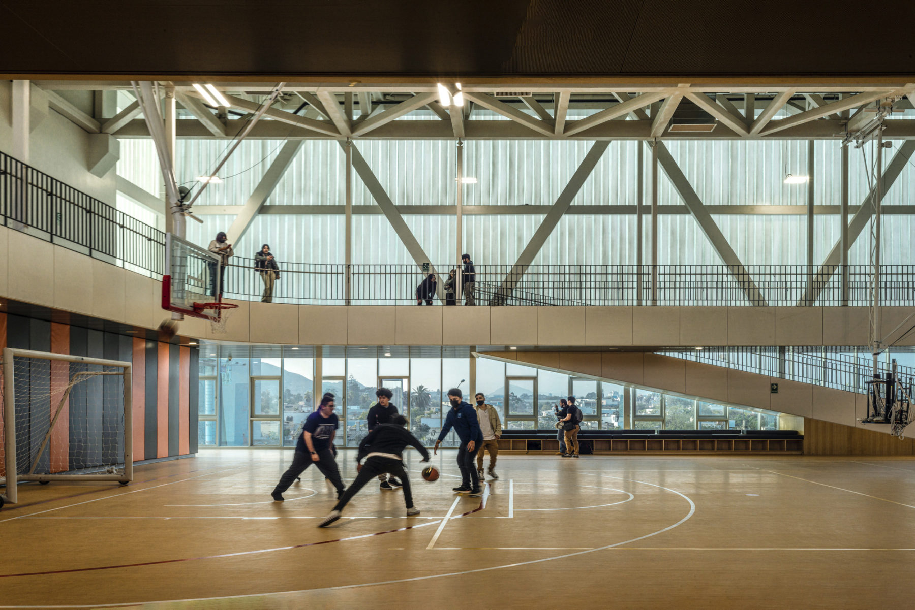 interior photo of gymnasium; a group of students is playing basketball in the foreground, another lounges on a bench near the court, and a third is on a bridge overlooking the court.
