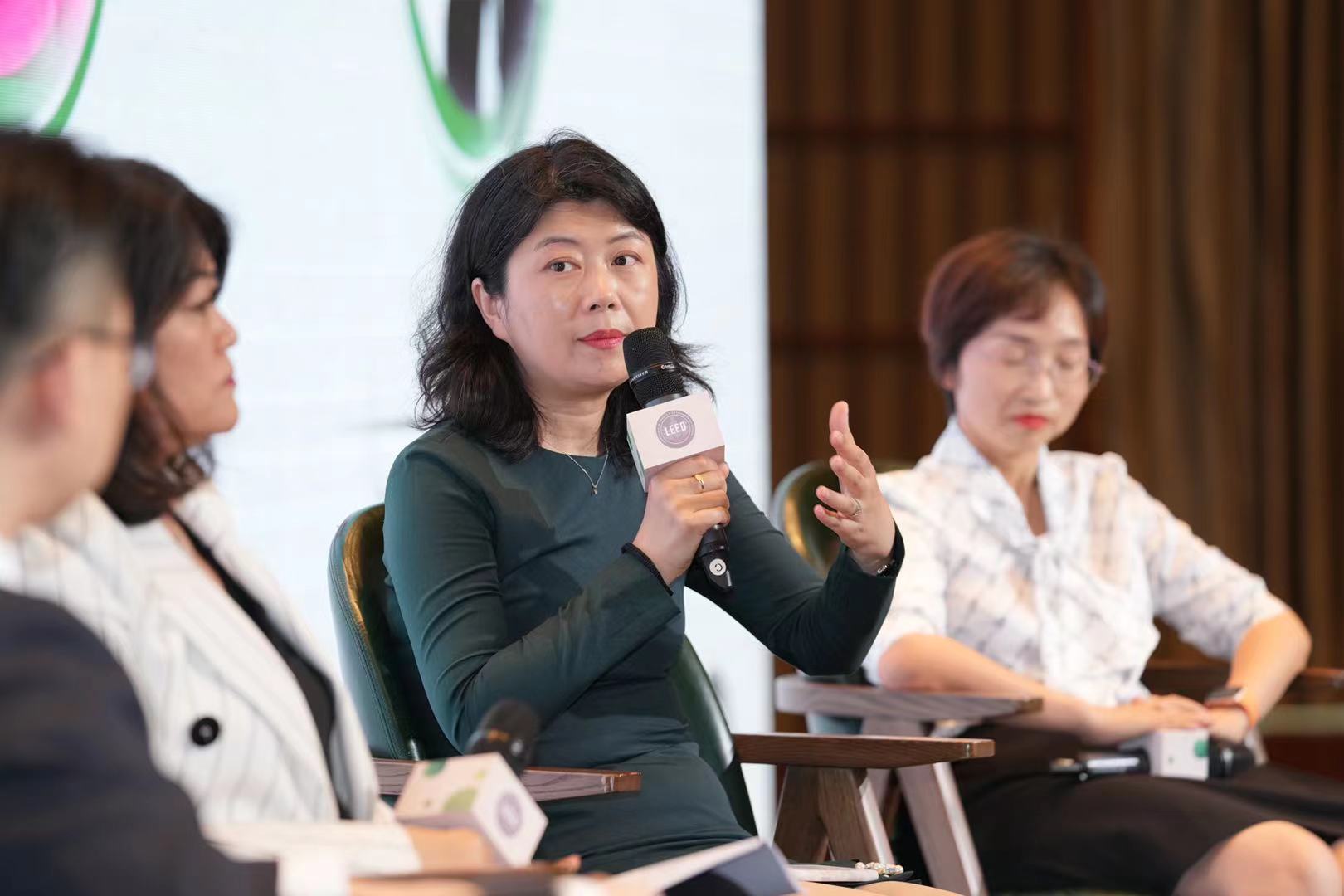 Dou Zhang sits on stage with three other panelists and a microphone in her hand, speaking