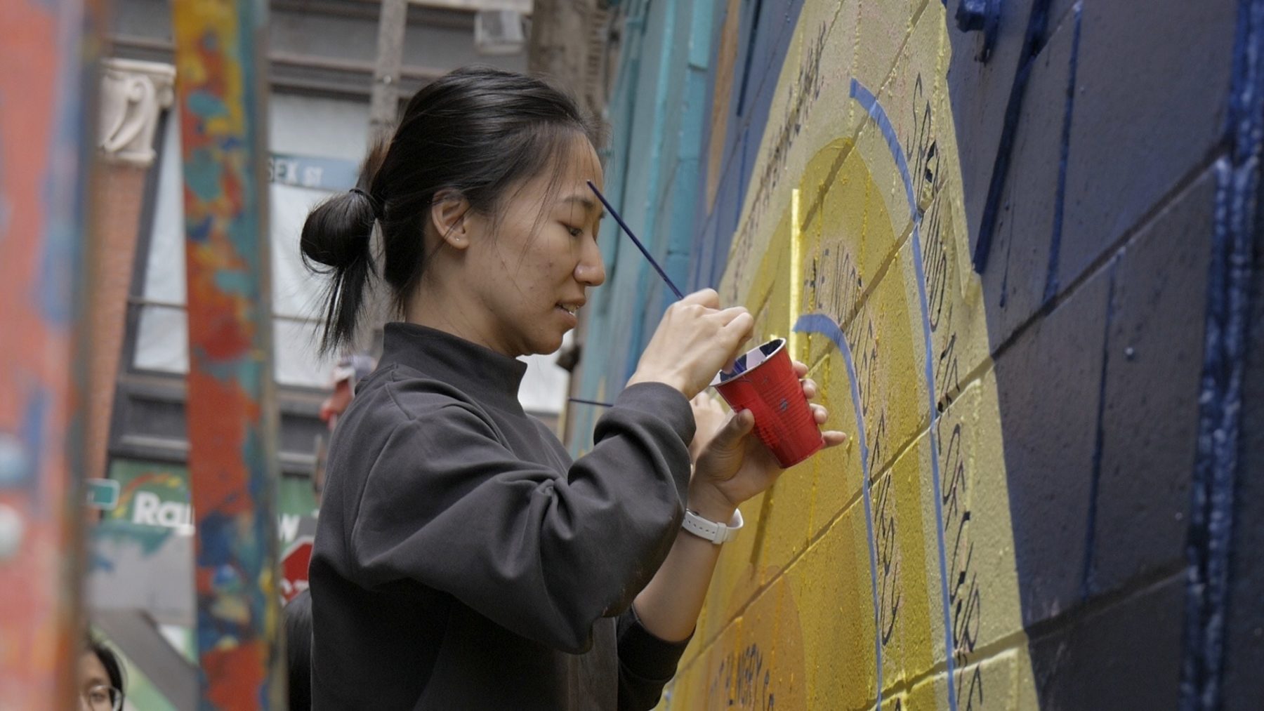 Volunteer dips paintbrush in cup of paint while helping with Chinatown mural