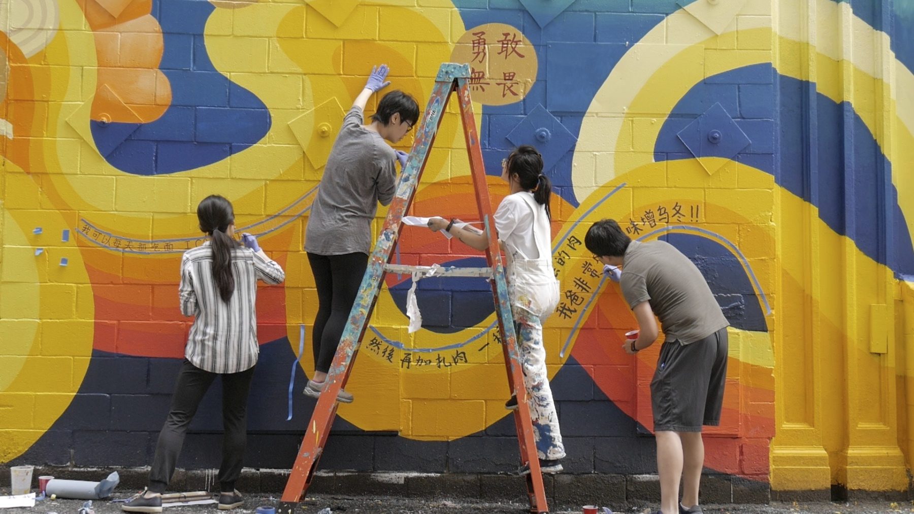 four people are shown standing on the ground and on a ladderpainting a mural