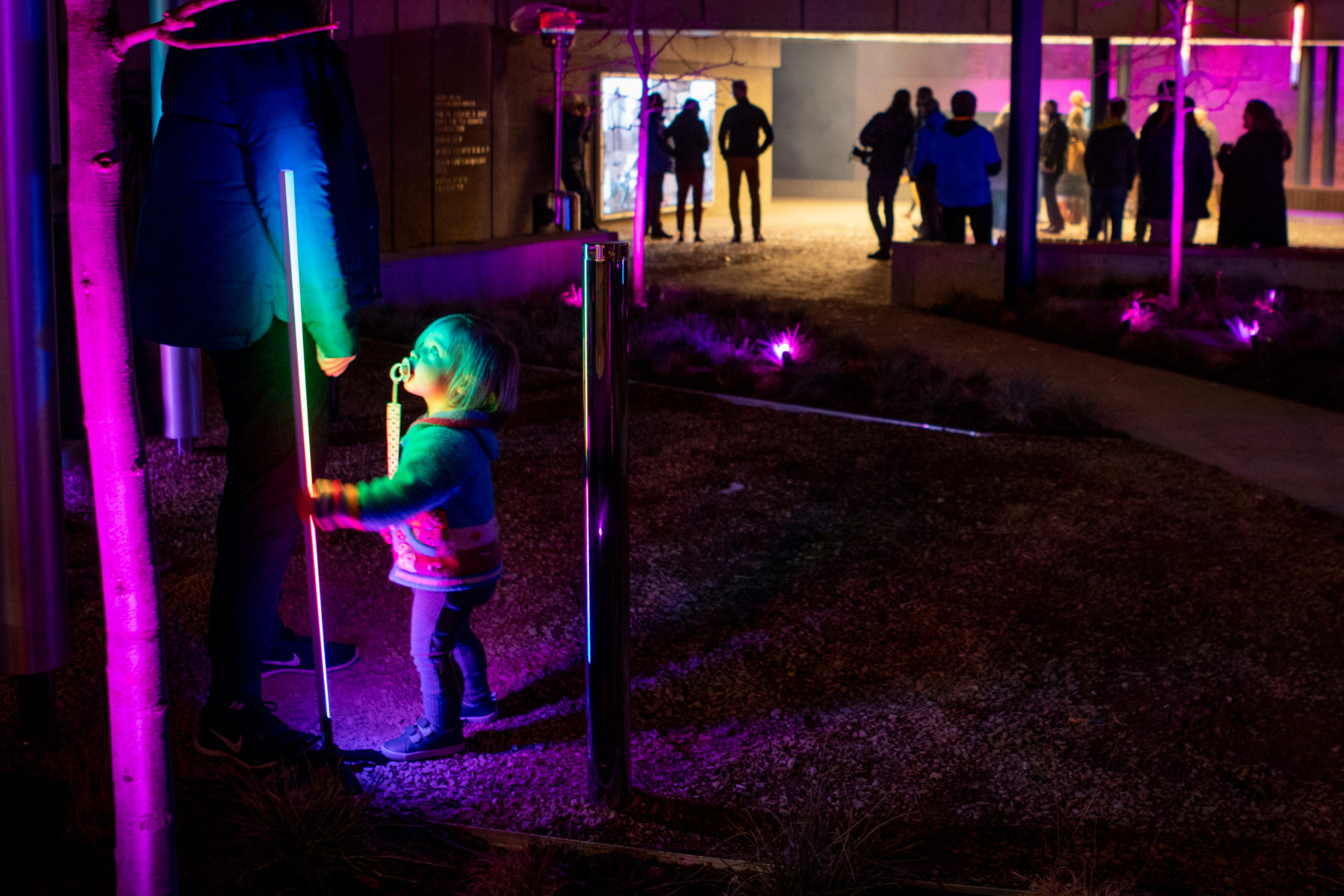 A toddler looks up at a pole installed in the plaza. The pole is illuminated and shines on the toddler's face