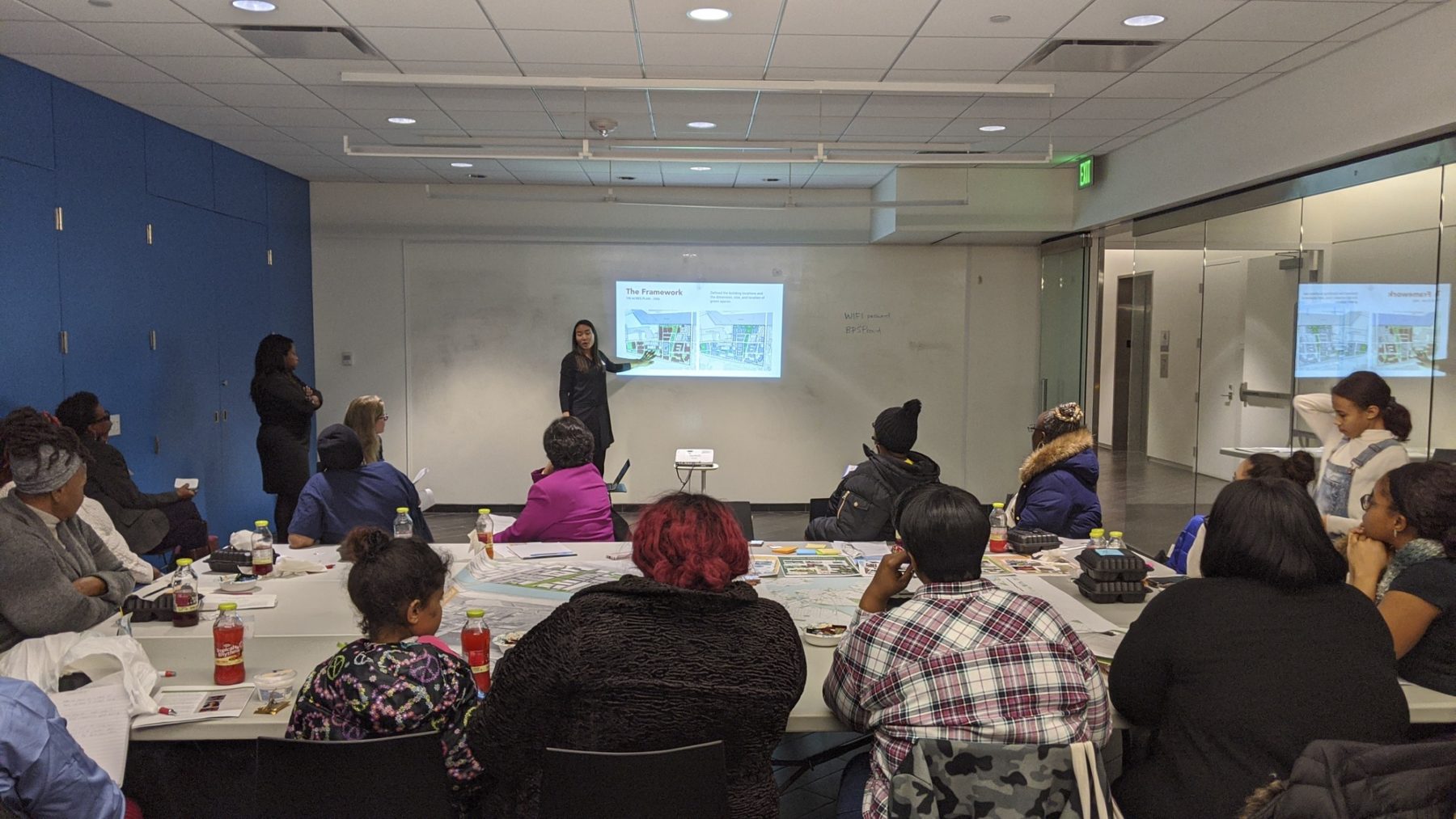 photo of public engagement session. Participants sit at a table a presenter points to a projection on the wall