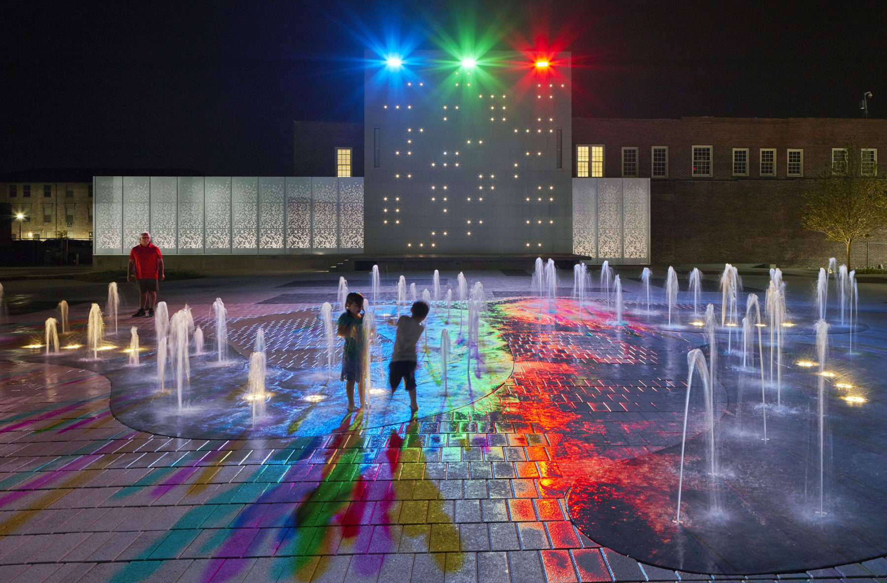 photo looking at video wall at night. in the foreground two kids play in a water feature