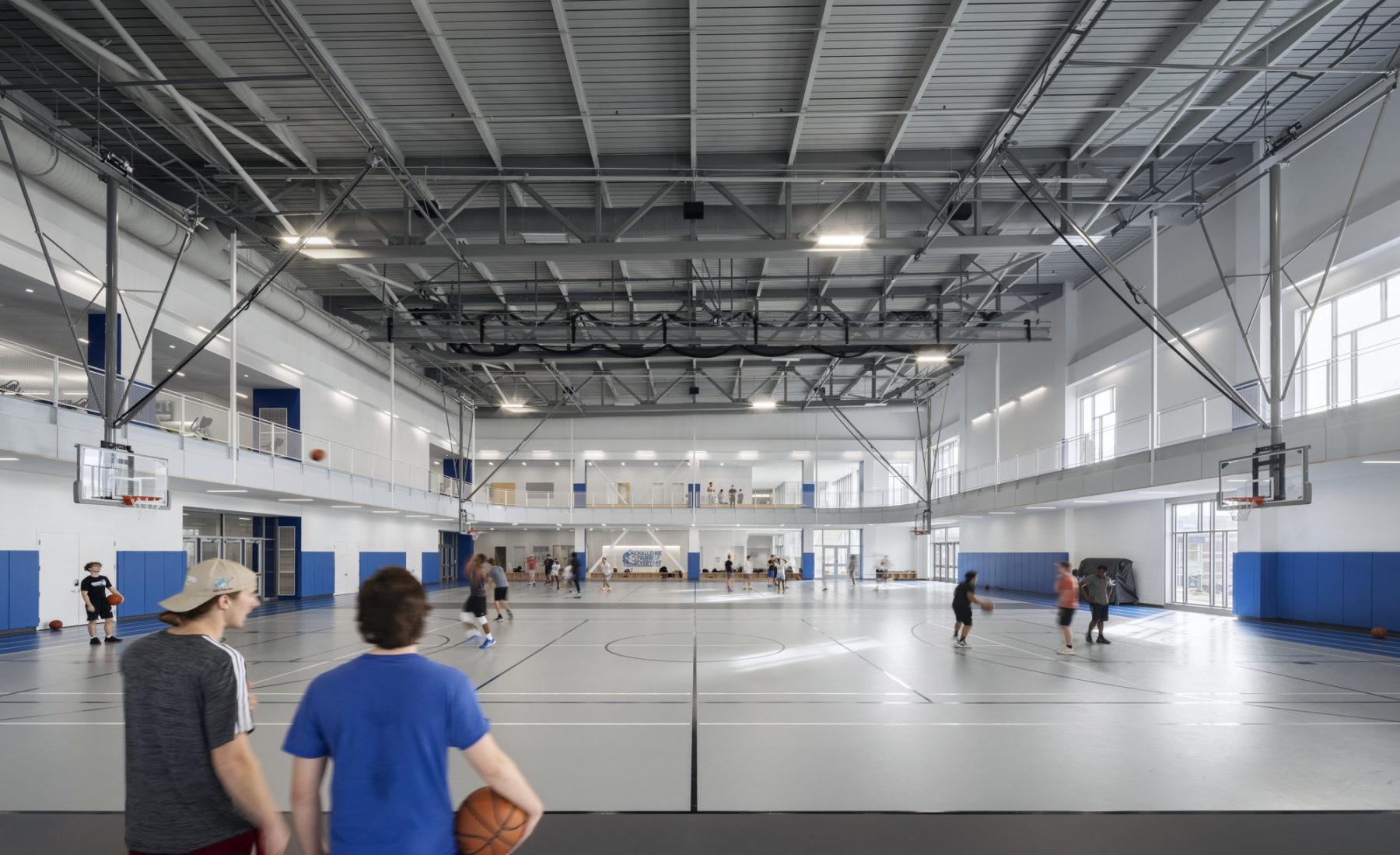 interior photo looking straight at basketball courts. A boy in a blue shirt looks at courts while holding a basketball in his right hand