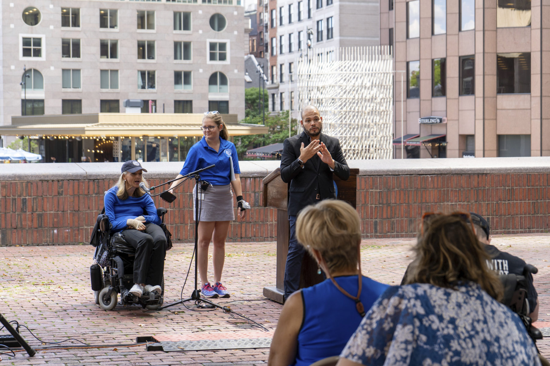 Woman in wheelchair speaking at event podium accompanied by ASL translator