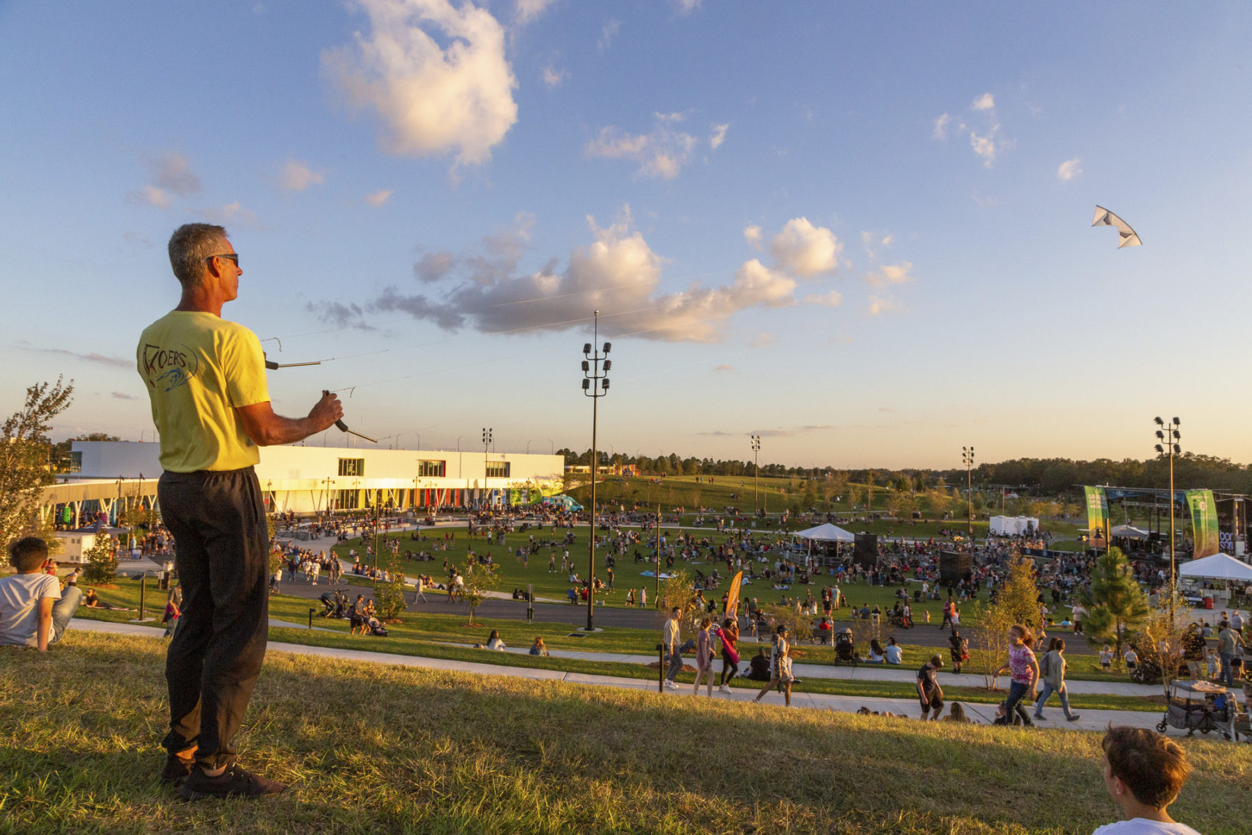 A man stands atop a hill in the foreground flying a kite at dusk. Below him, the park slopes downward and is full of people and activity