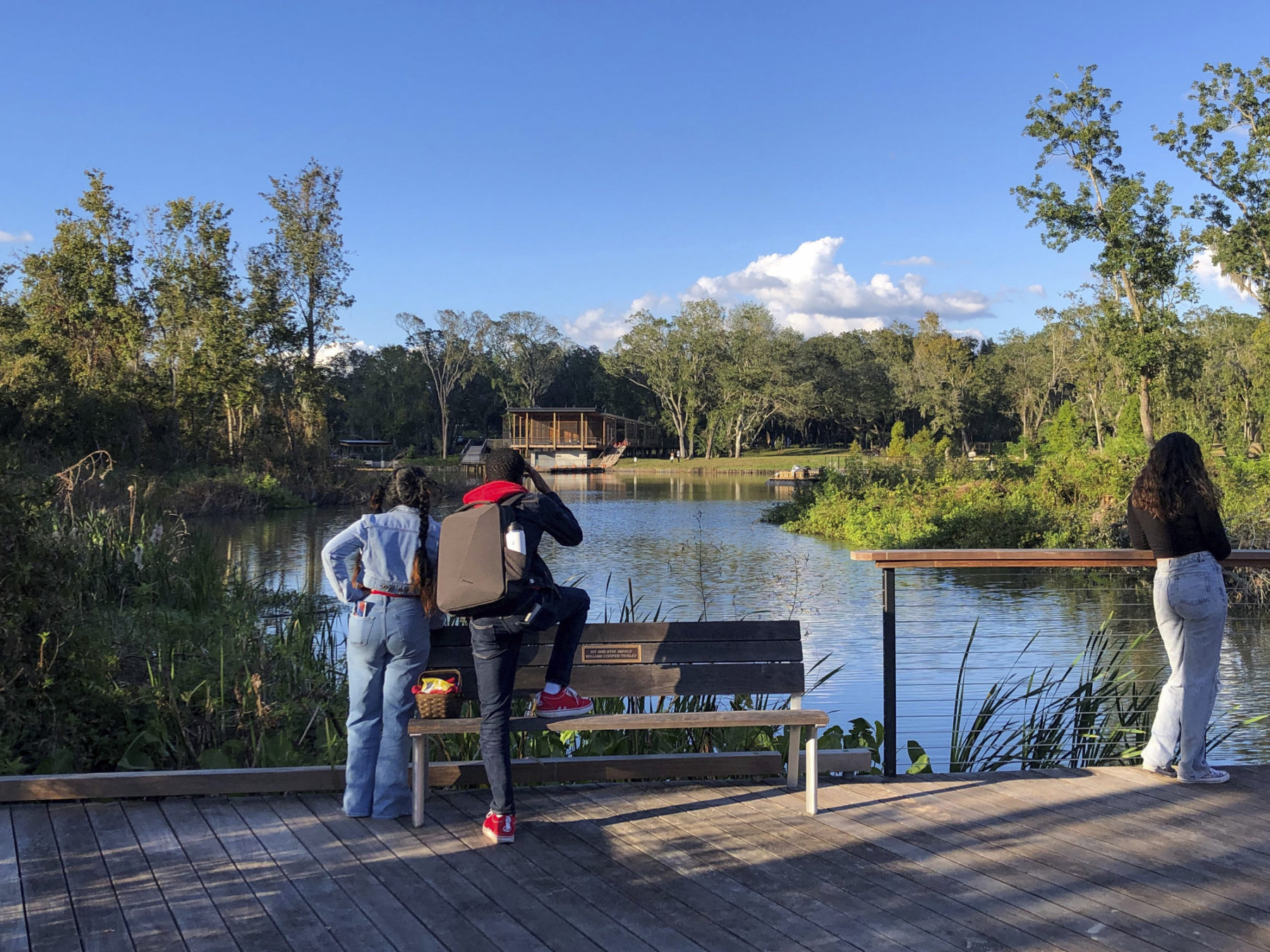Two people stand by a bench overlooking a lagoon on a sunny day. In the distance a wooden building is visble.