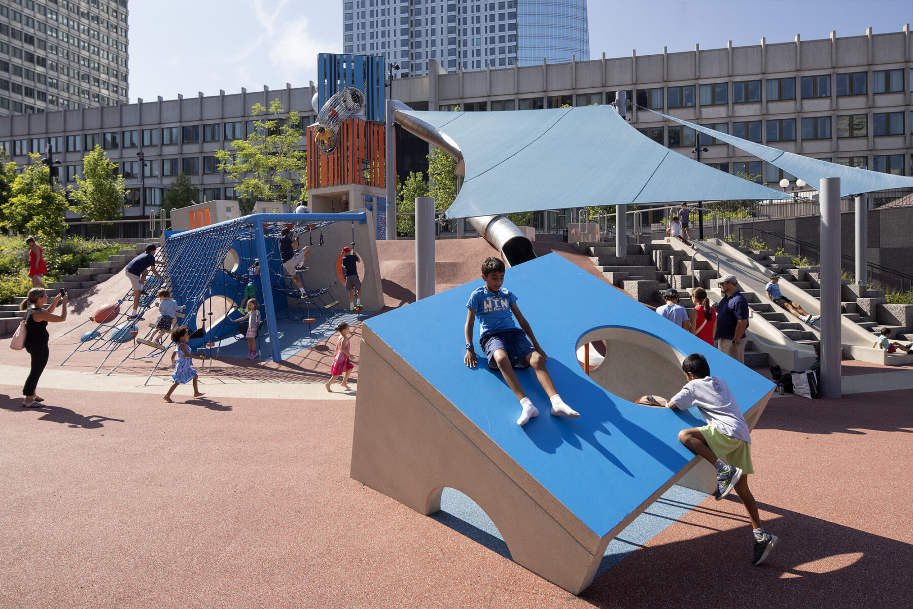 Two boys climbing on cube play structure in foreground of the play space with children running and climbing on play equipment in background
