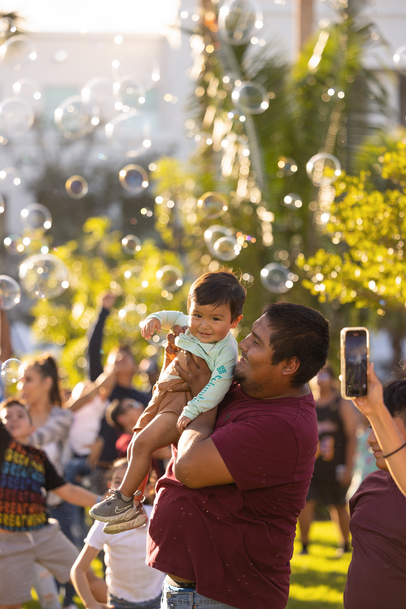 A man holding up a toddler in a crowd of people as someone blows bubbles around them