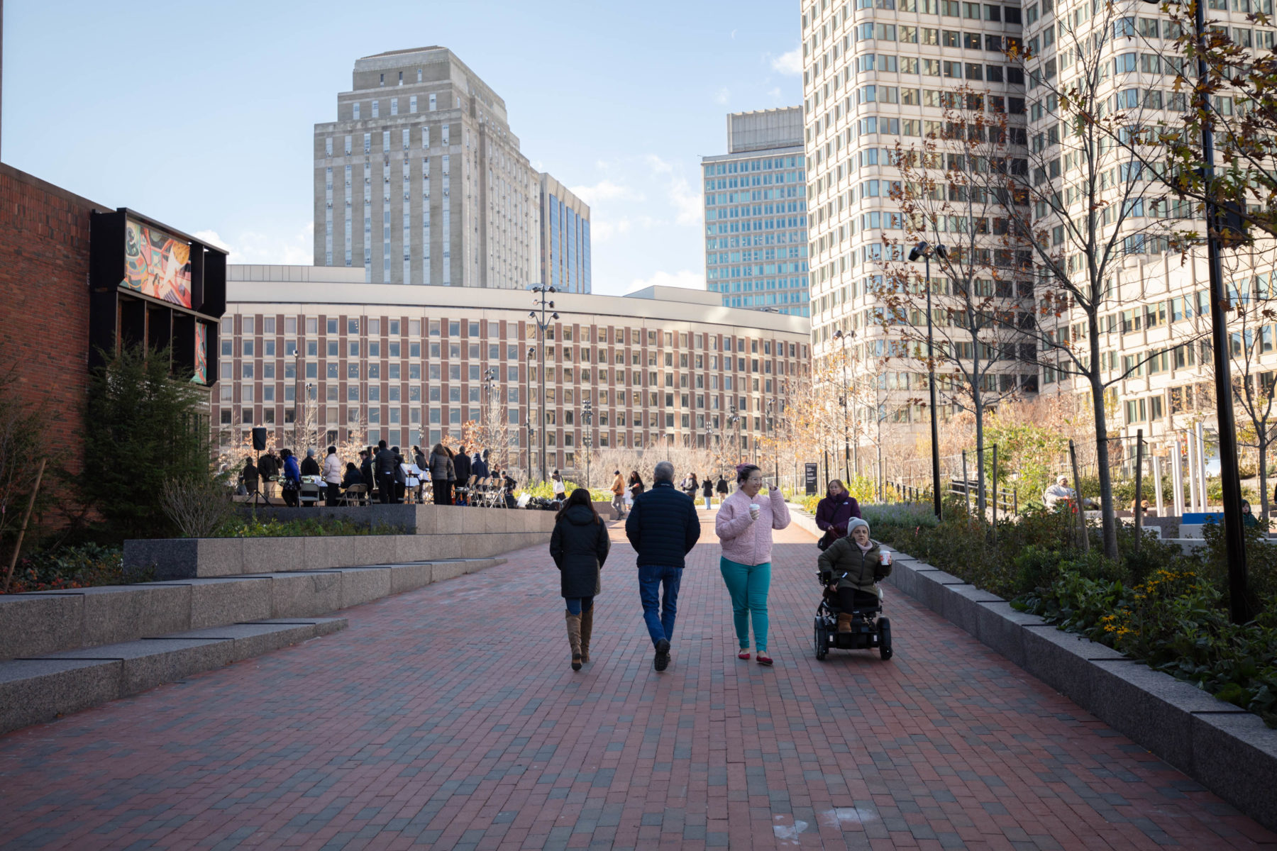 Four people moving down a brick pathway. Three people are walking and one person is using an electric wheelchair.