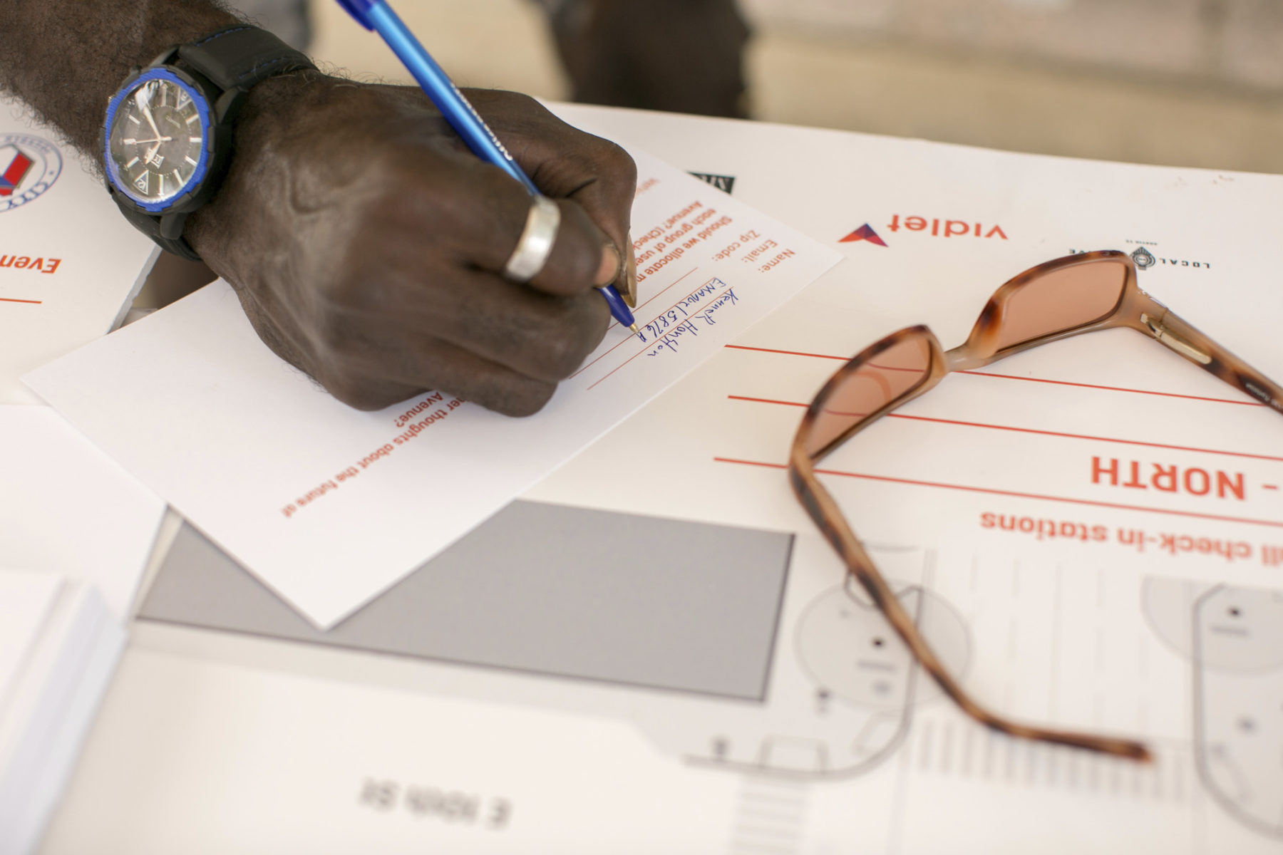 A detail photo of a resident's hand filling out a comment card at a public engagement session. Sunglasses sit on top of a stack of cards.