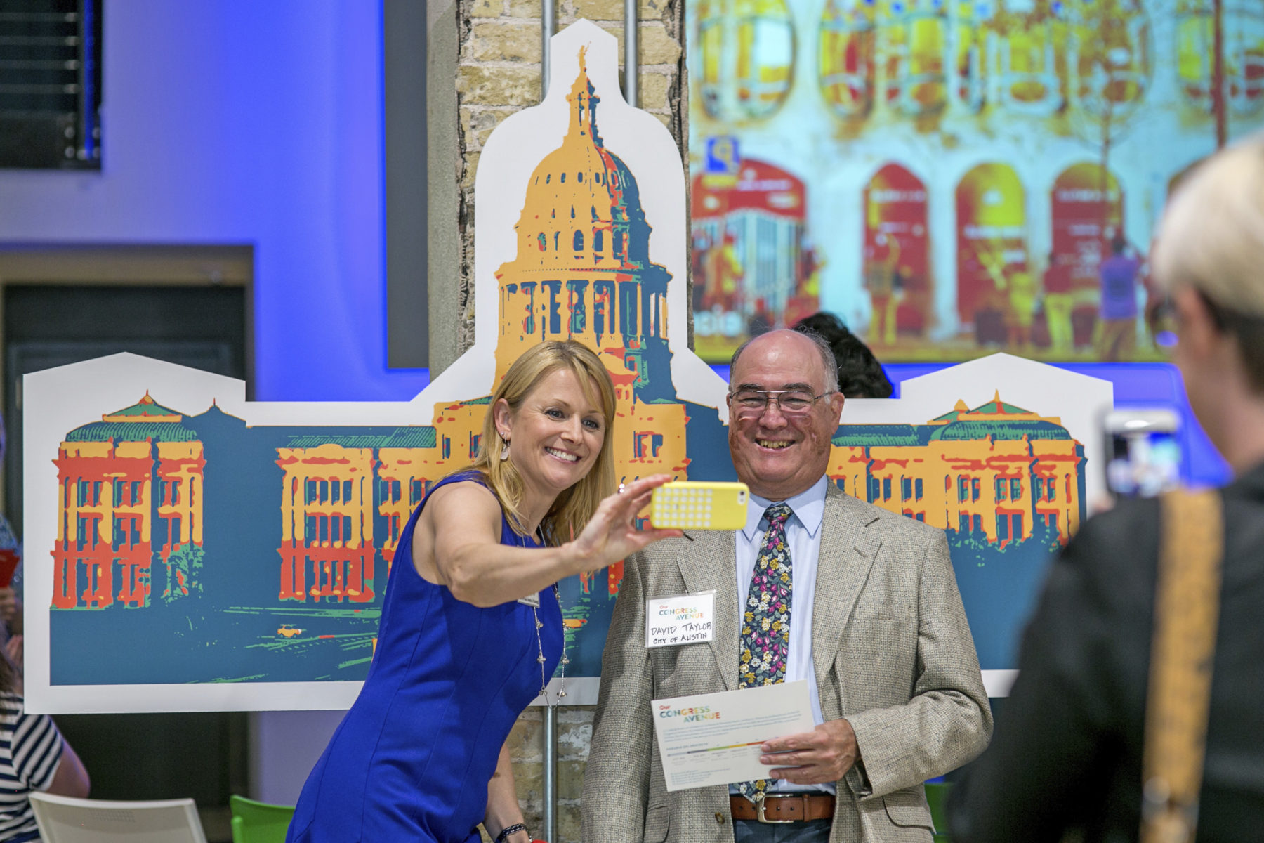 photo of a woman in a blue dress taking a selfie on a yellow phone with a bald man in a suit jacket and tie. They are standing in front of a rendered print out of the Texas State Capitol building.