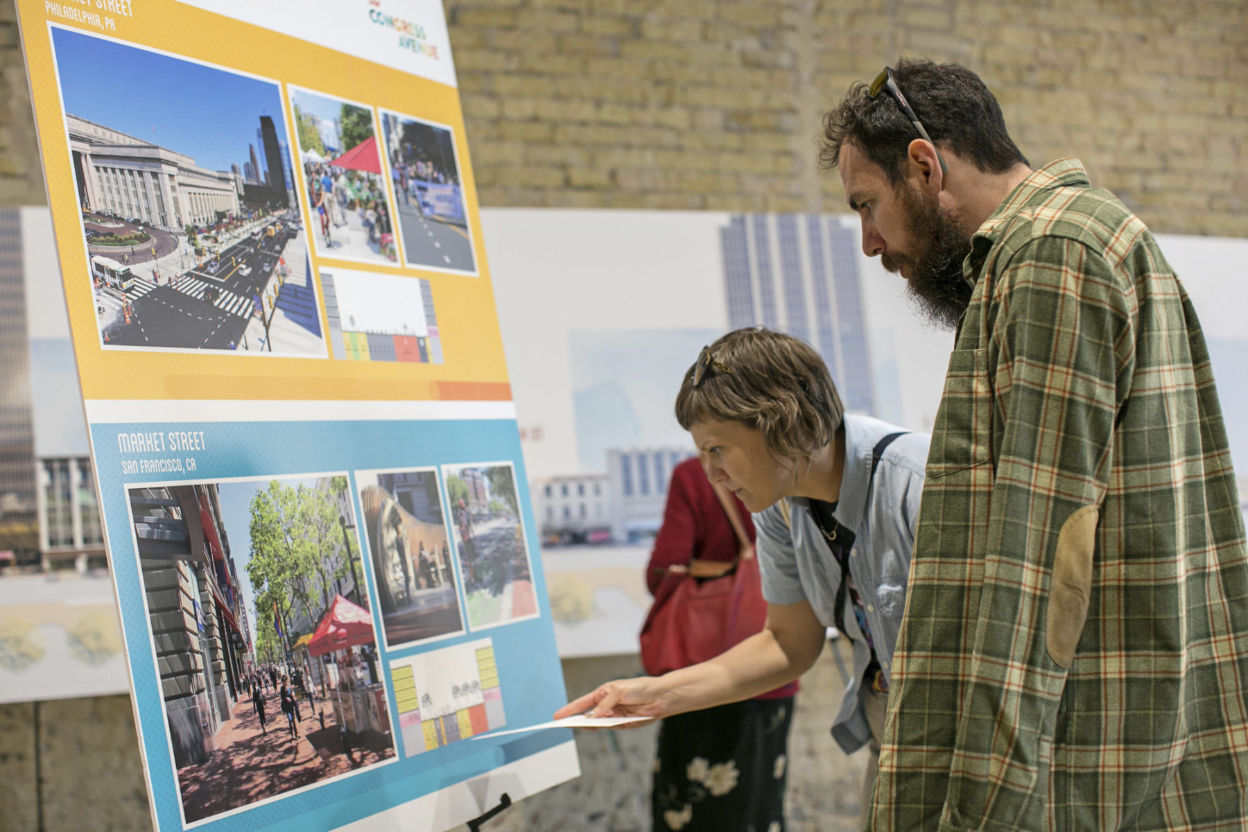 Photo of a man and a woman looking at a board that has pictures and sections of similar streets in other major cities. The woman is crouched down and pointing to a street section and the man looks down at it. He is wearing a green plaid shirt.