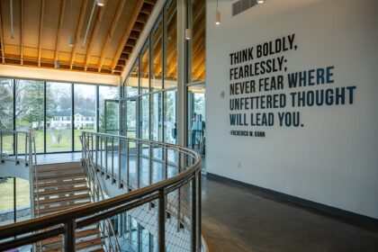 Interior photograph with wall graphics, a staircase and landing, and glass windows with views to campus