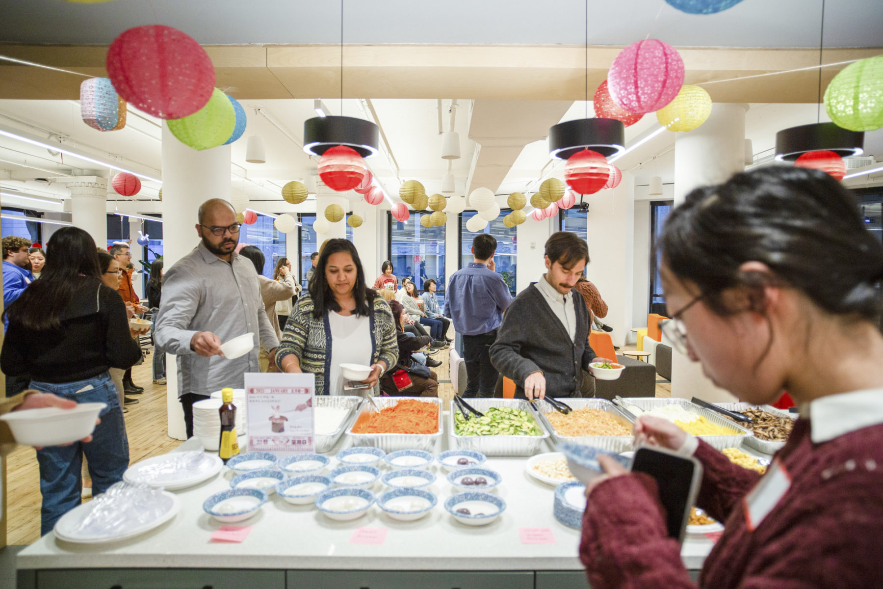 People getting food in the foreground with other people milling about in the background. Lanterns hang from the ceiling