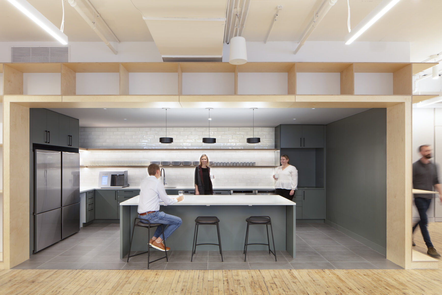 people conversing in an open kitchen with overhead shelving and person walking by in background