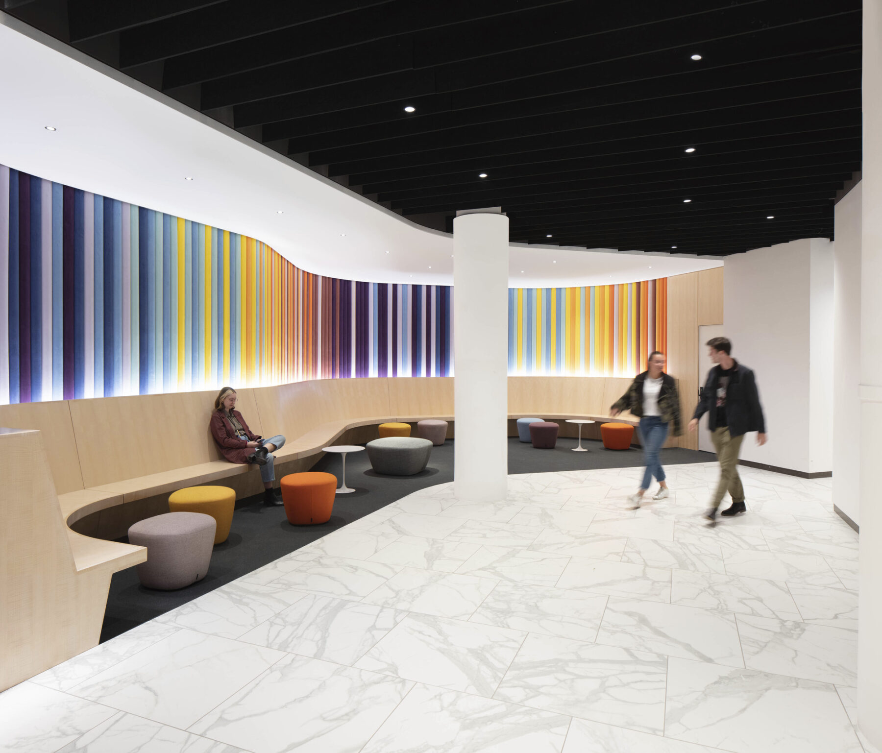Lobby space with wrap around built-in bench and colorfully upholstered wall. Two people walk through the space chatting while one person sits on the bench looking at her phone.