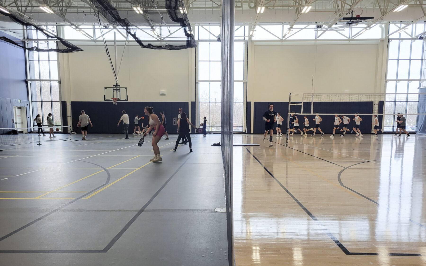 A gym divided in two by a screen. On the left side there is a blue floor and students play pickleball. On the right side there is a wood floor and students are jogging in a group