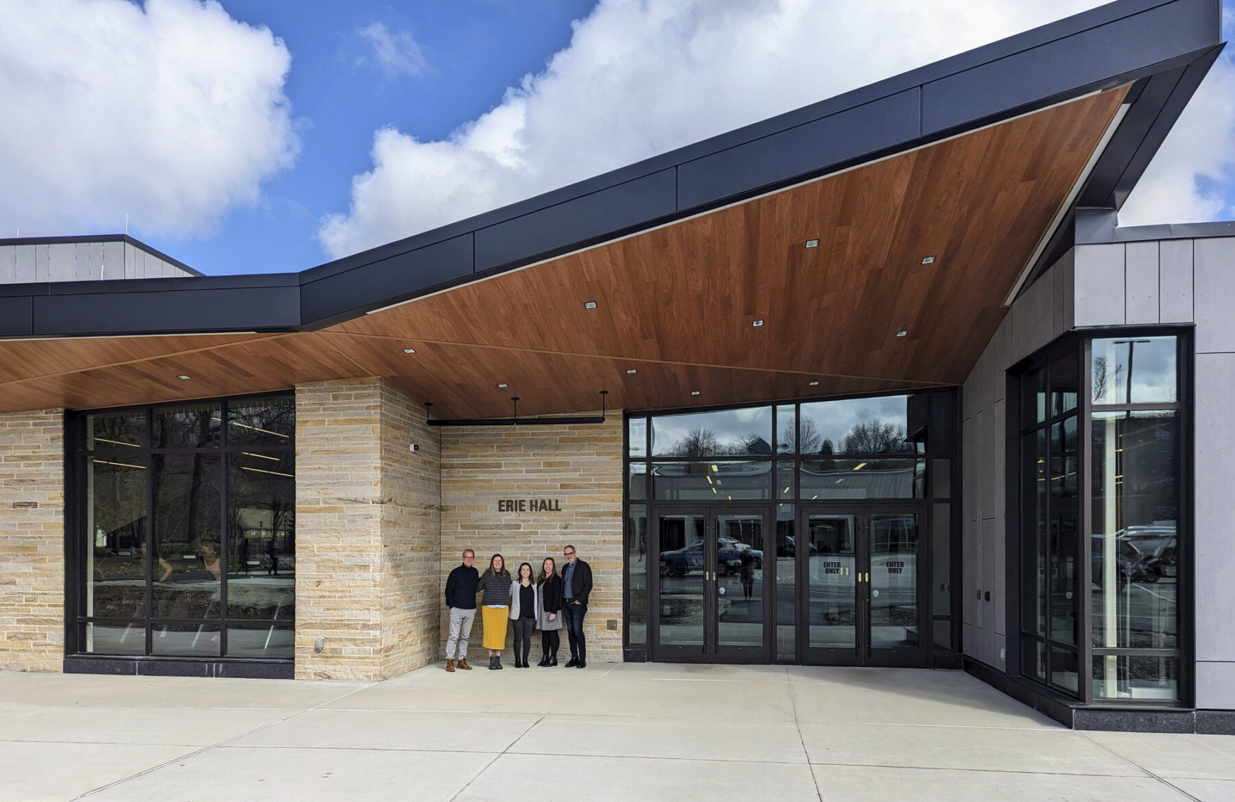 Four people smiling, posing in front of building with an angular overhang. blue sky above them