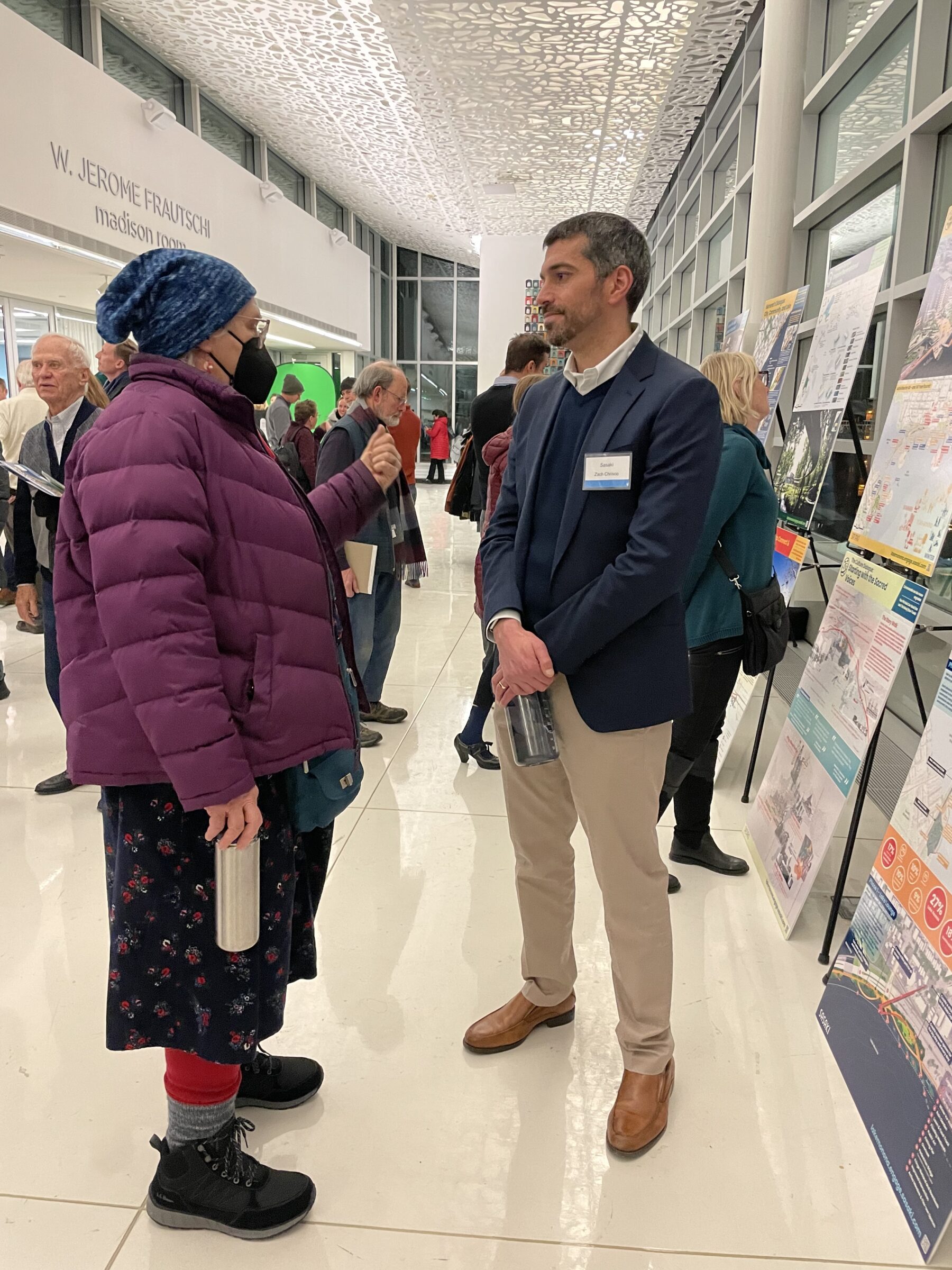 sasaki principal zachary chrisco, a tall man in a suit jacket, talks to a community member who is wearing a mask and a purple jacket. they are indoors at a community engagement event