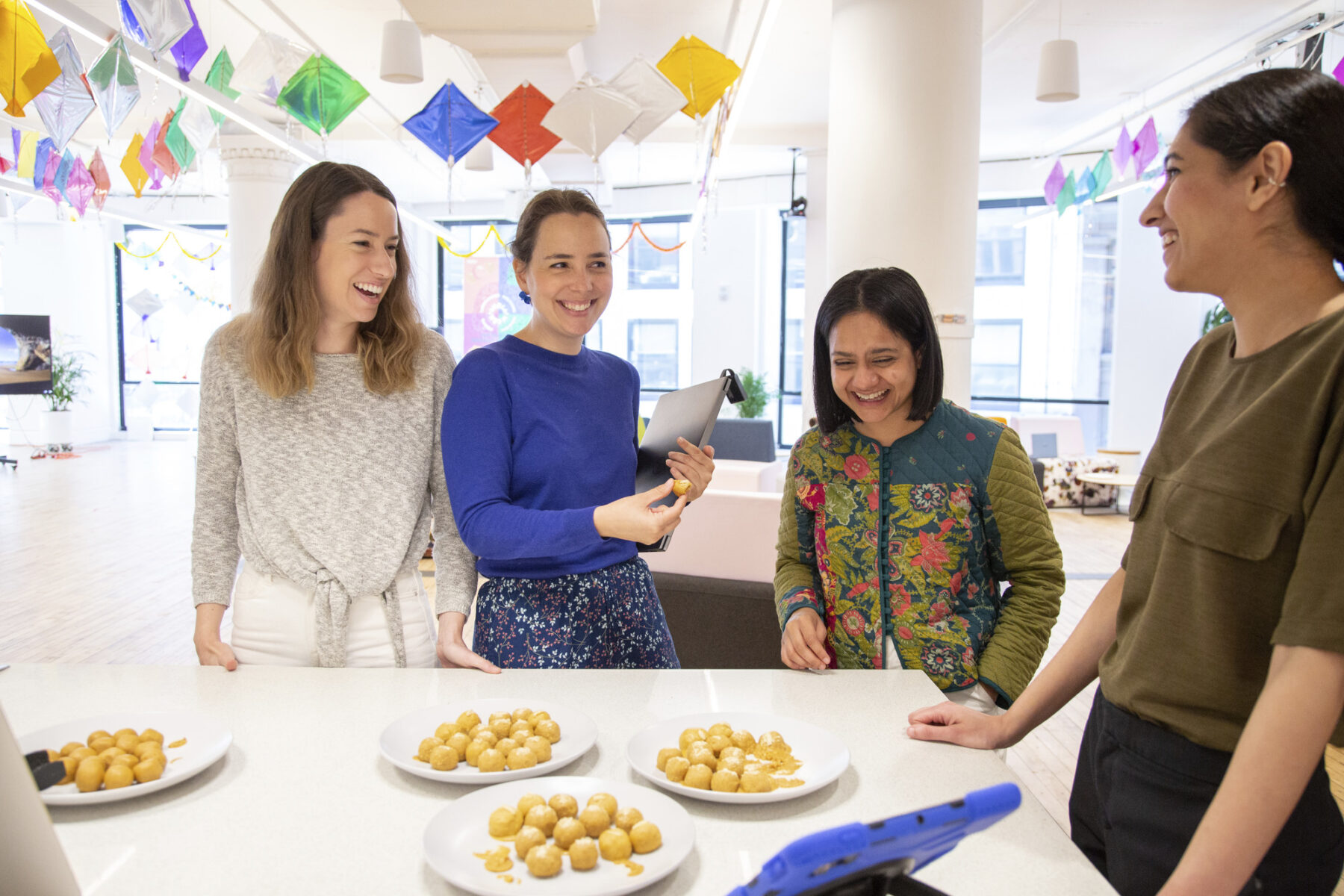 a photo of people gathered around a kitchen counter smiling. There are plates of laddoo on the table.