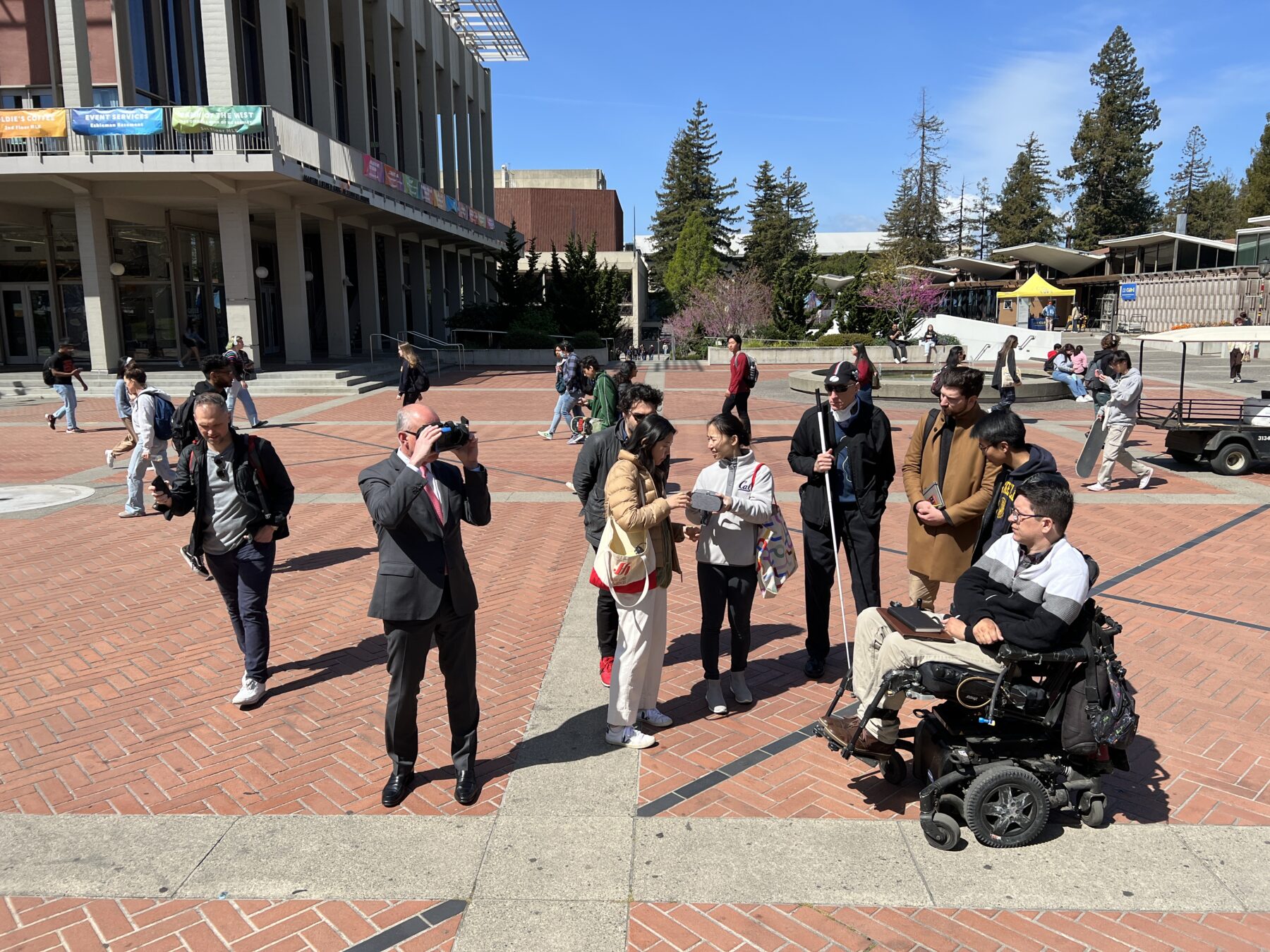 people gathered on a brick plaza. one person uses a VR headset. Several people are standing and one is in a wheelchair