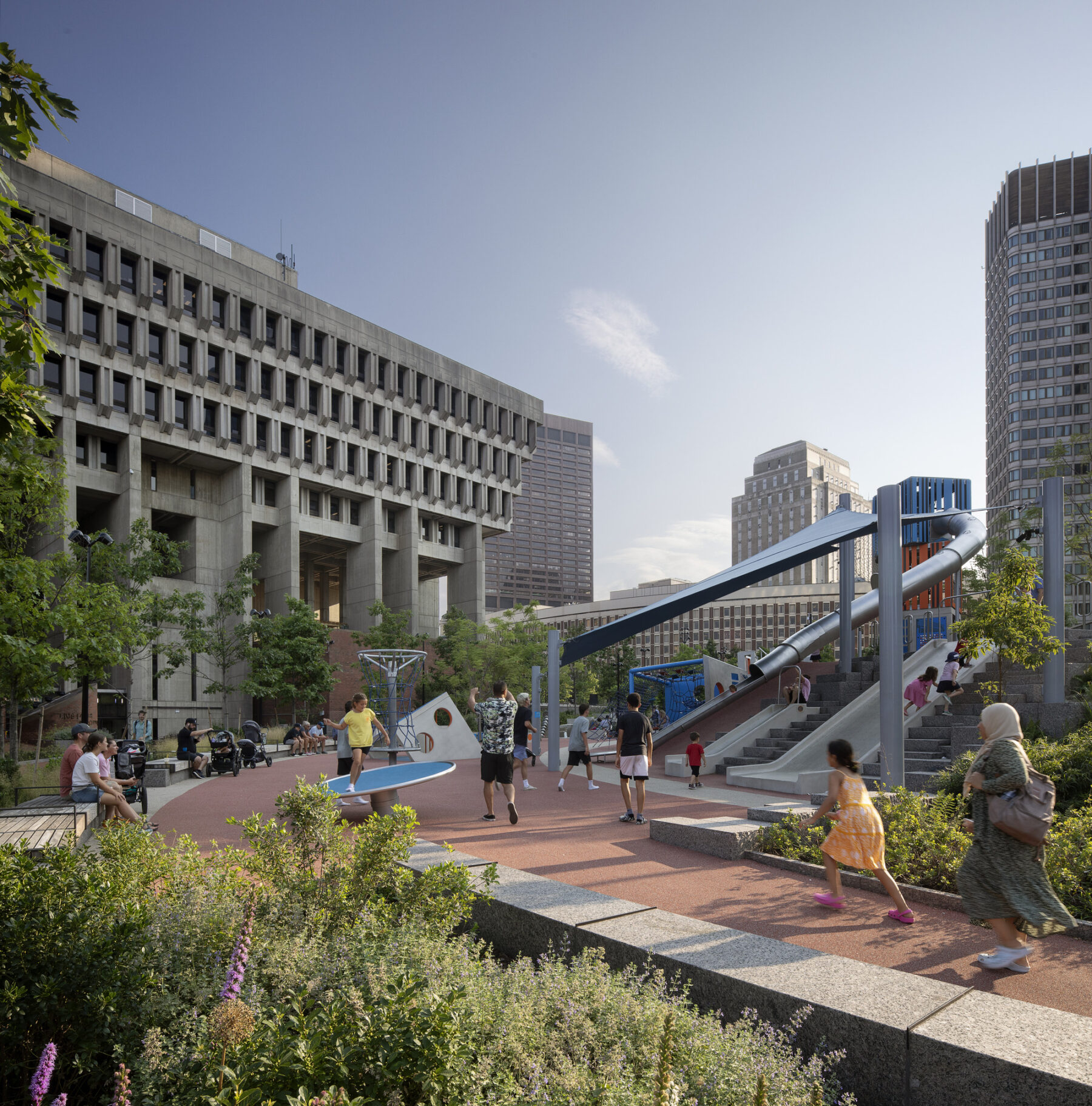 Afternoon photo: vegetation in the foreground, children slide and frolic, and parents watch, at the playscape adjacent to Boston City Hall