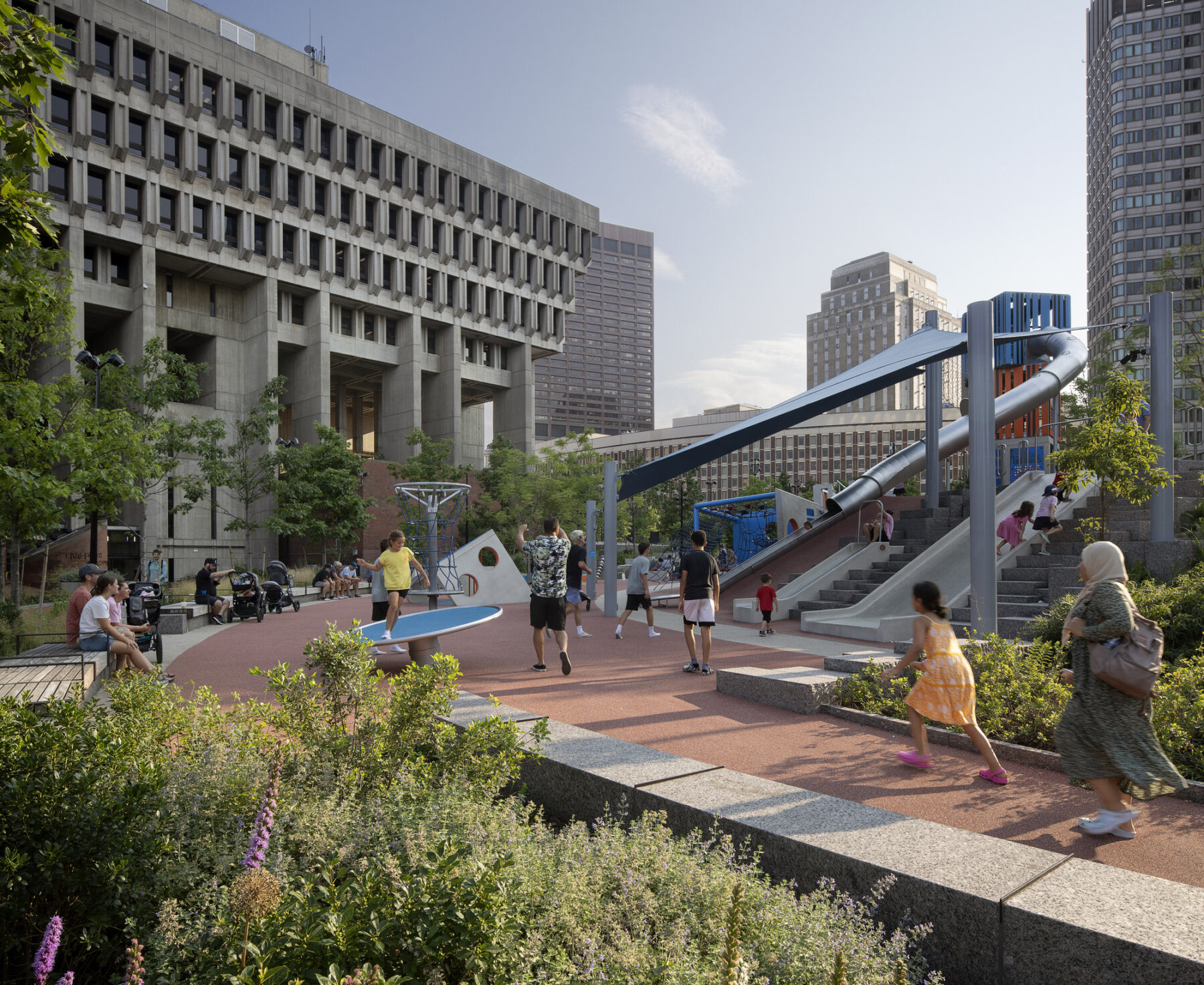 Afternoon photo: vegetation in the foreground, children slide and frolic, and parents watch, at the playscape adjacent to Boston City Hall