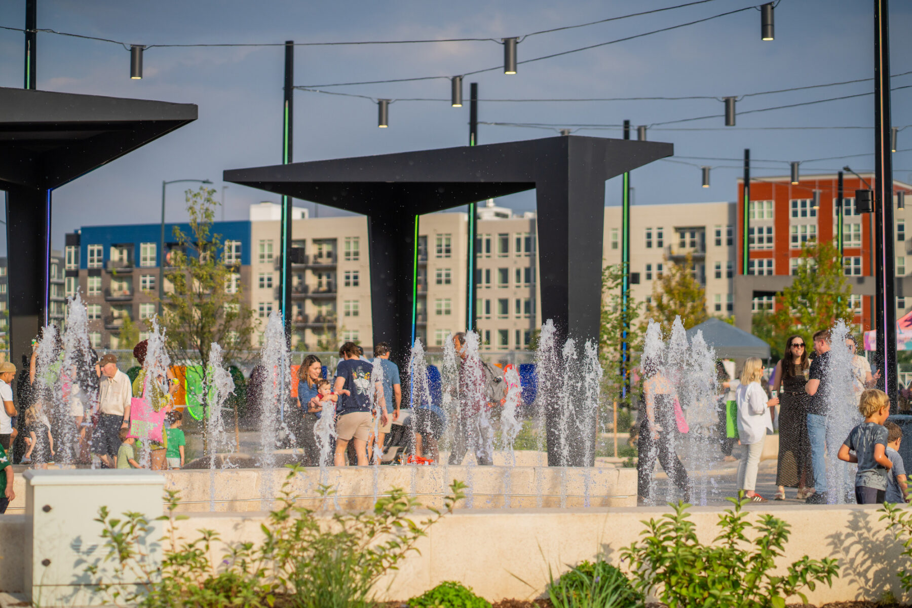 A view across the plaza with strung light fixtures, black metal pergola structures, water fountains in the foreground, and visitors enjoying the amenities.